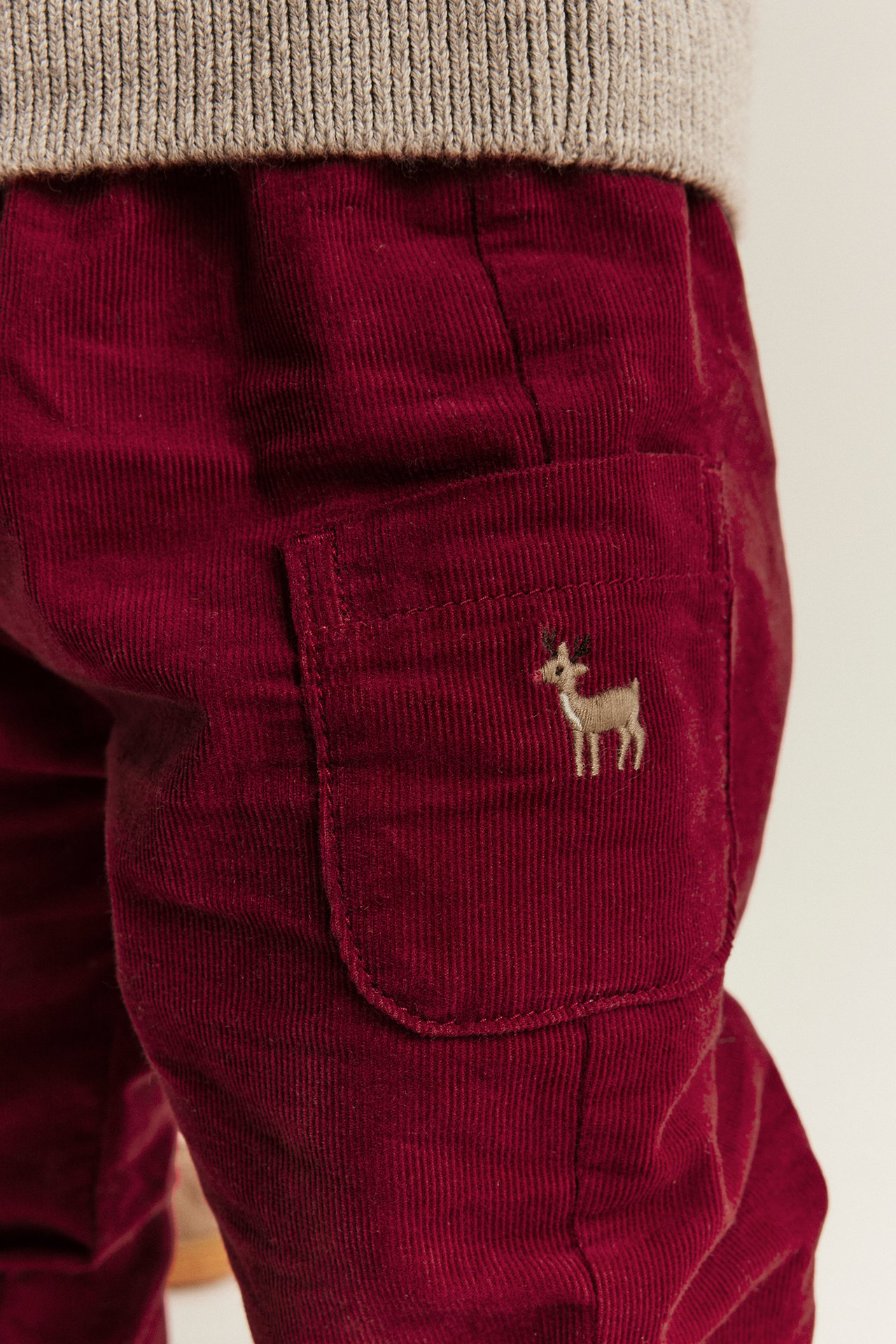 Close-up of kid's red corduroy pants with a deer embroidery and a beige knitted sweater.