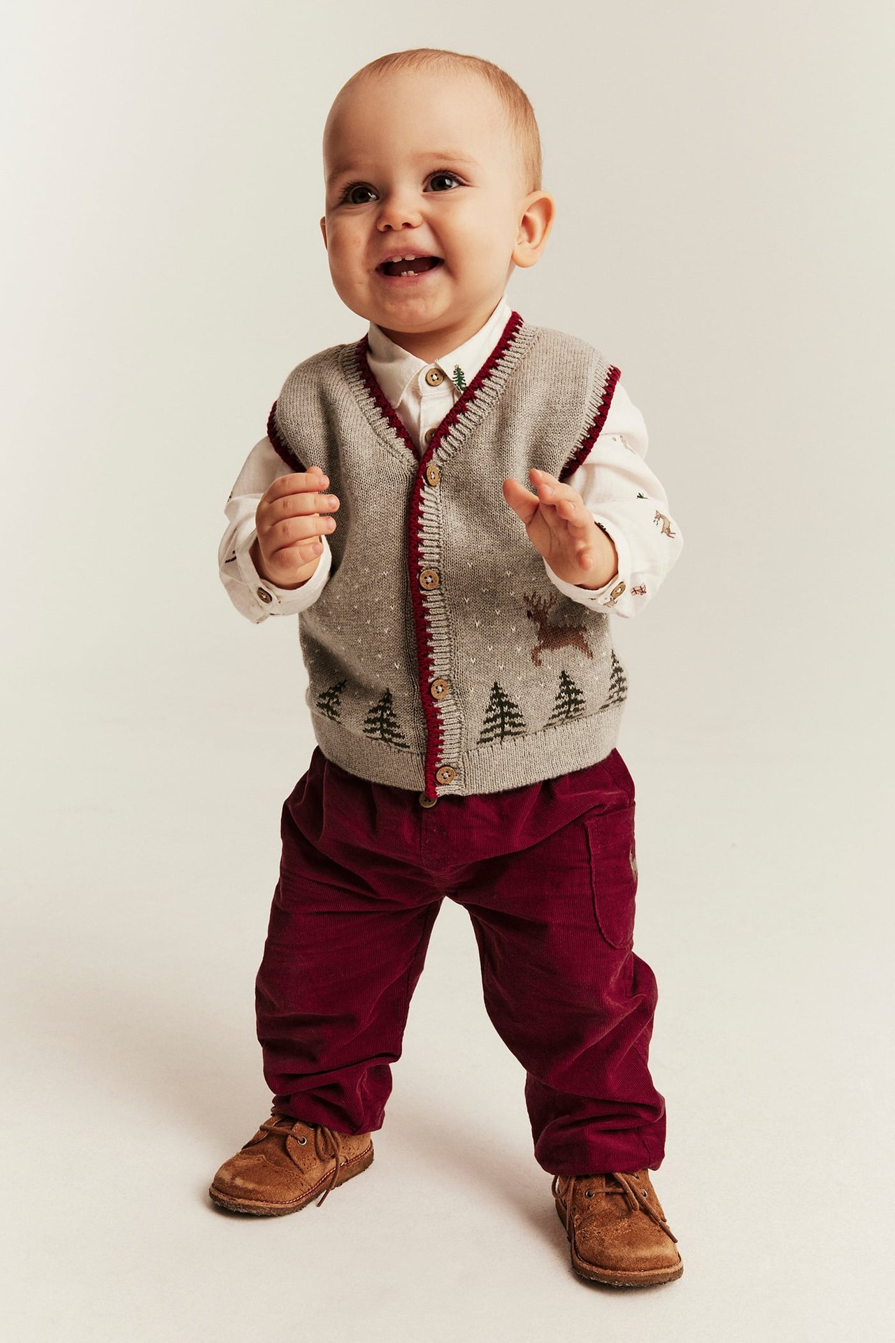 Vorderansicht Baby in grauer Strickweste mit Hirsch und Bäumen, weißem Hemd, rote Hose, braune Schuhe.