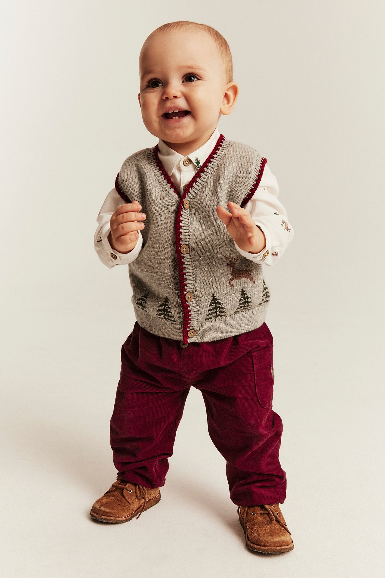 Vorderansicht Baby in grauer Strickweste mit Hirsch und Bäumen, weißem Hemd, rote Hose, braune Schuhe.