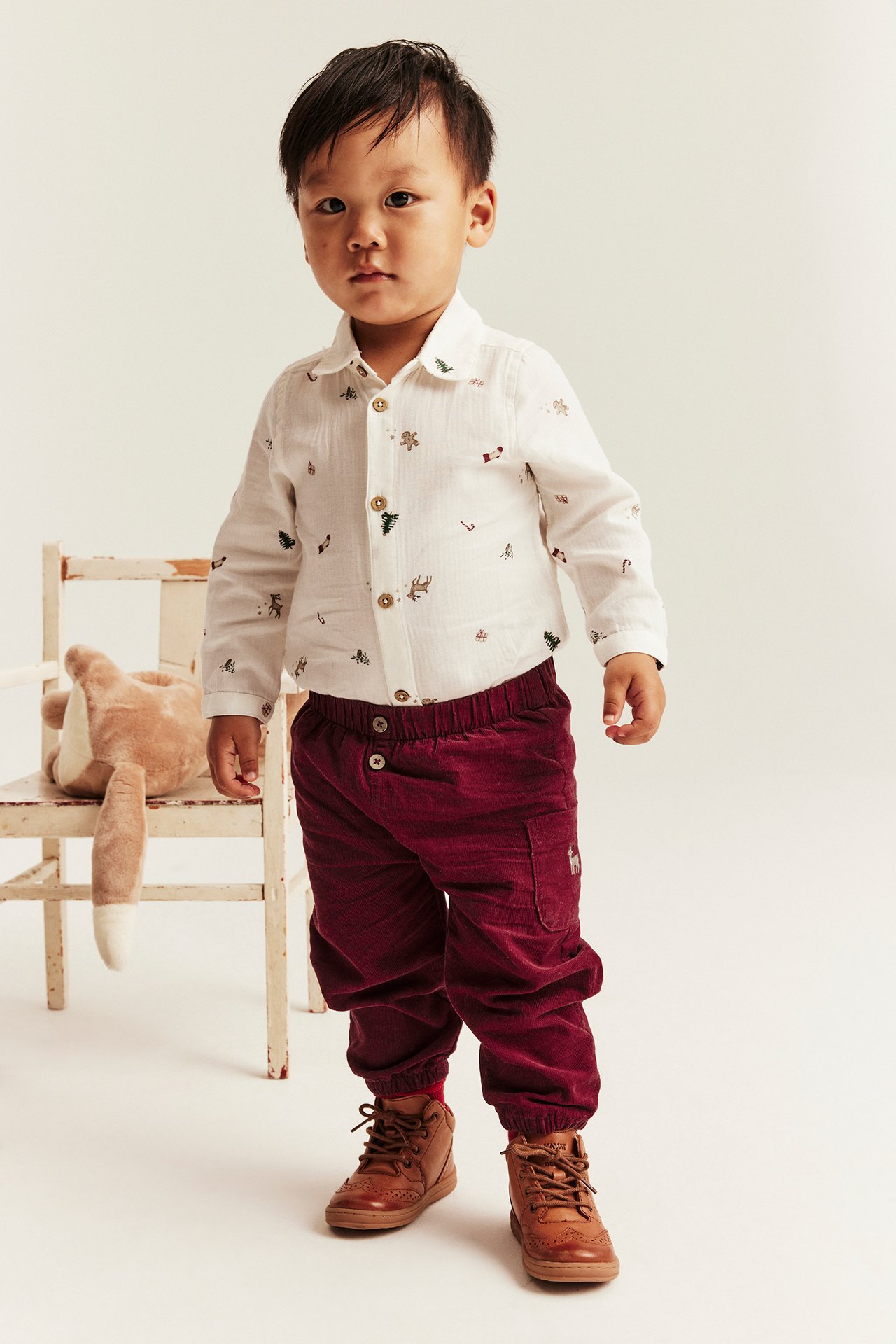Front view of a kid in a white festive print shirt, burgundy corduroy pants, and brown lace-up boots.