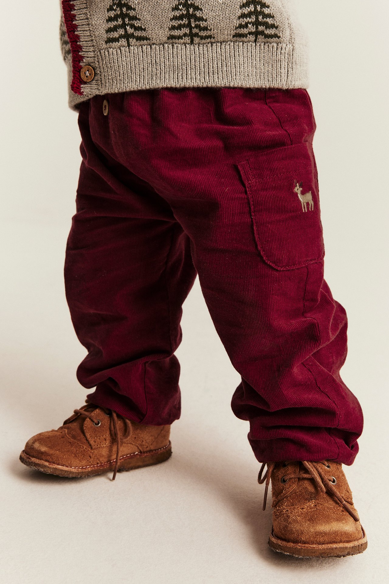 Kind in bordeauxroter Cordhose mit Hirsch, grauem Cardigan und braunen Schnürschuhen.