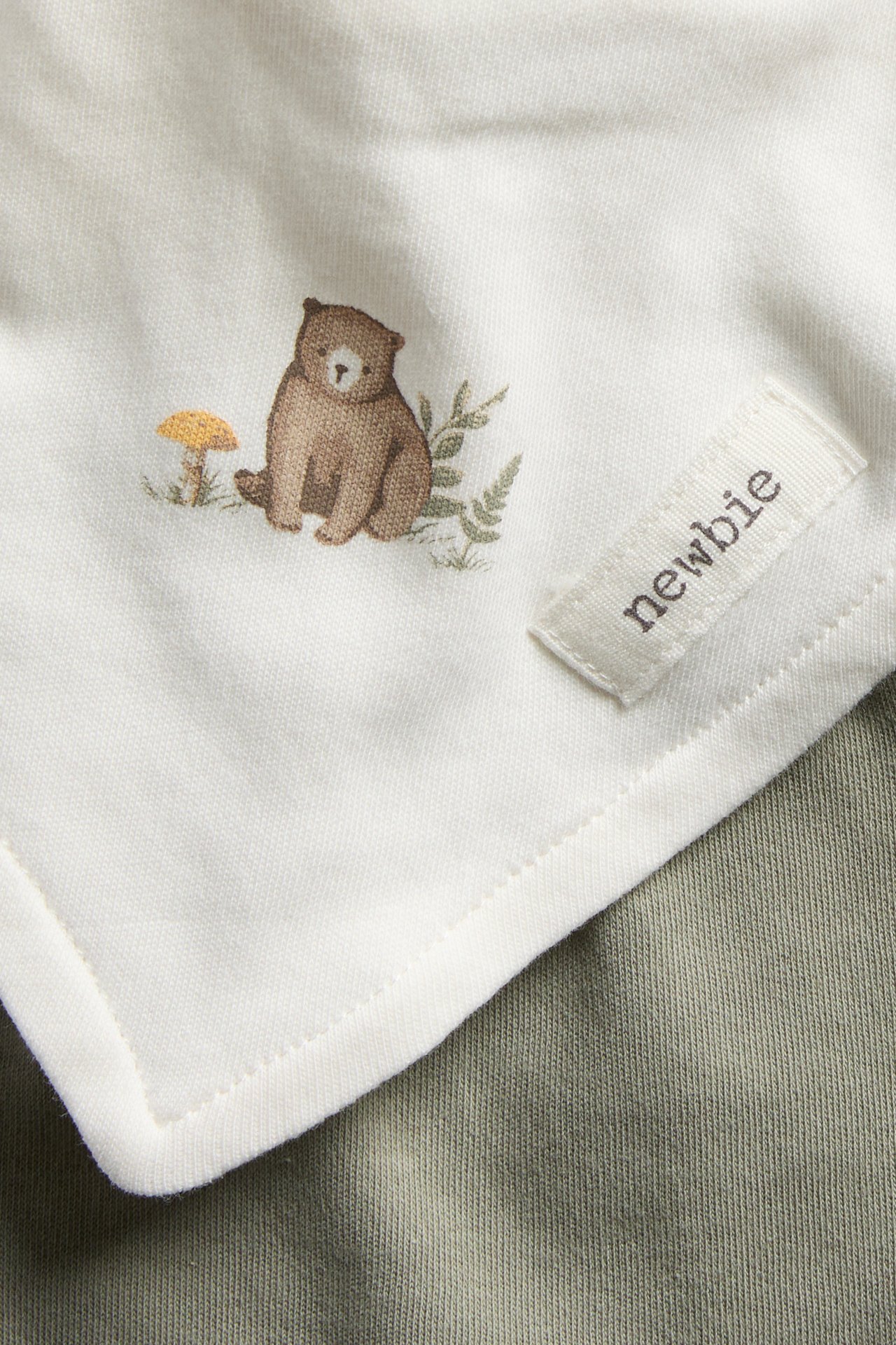 Close-up of an off-white cotton baby bib with a brown bear print and 'newbie' label.