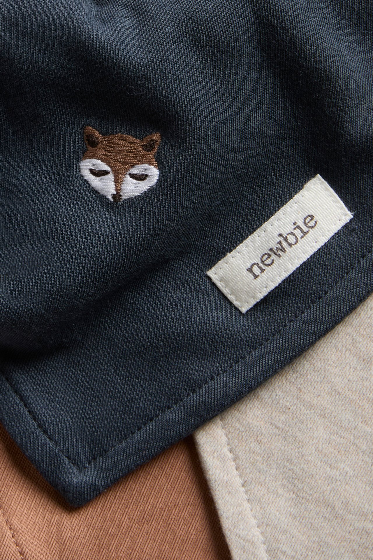 Close-up of a dark blue baby bib with brown and white fox embroidery and a 'newbie' tag.