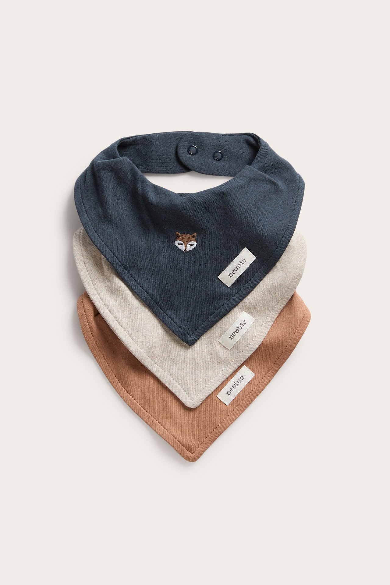 Front view of three stacked baby bandana bibs: dark blue with fox embroidery, beige, and brown.