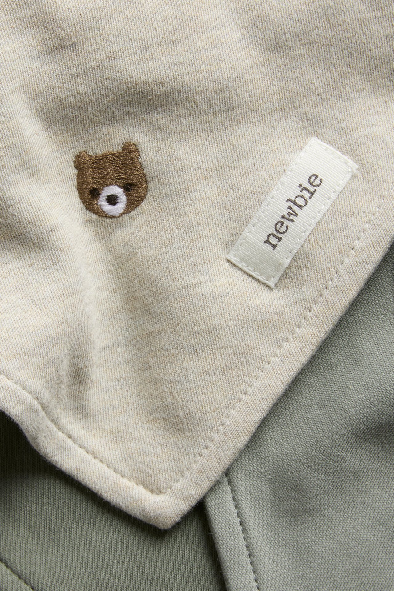 Close-up: light beige baby garment with embroidered bear and label, on green fabric.