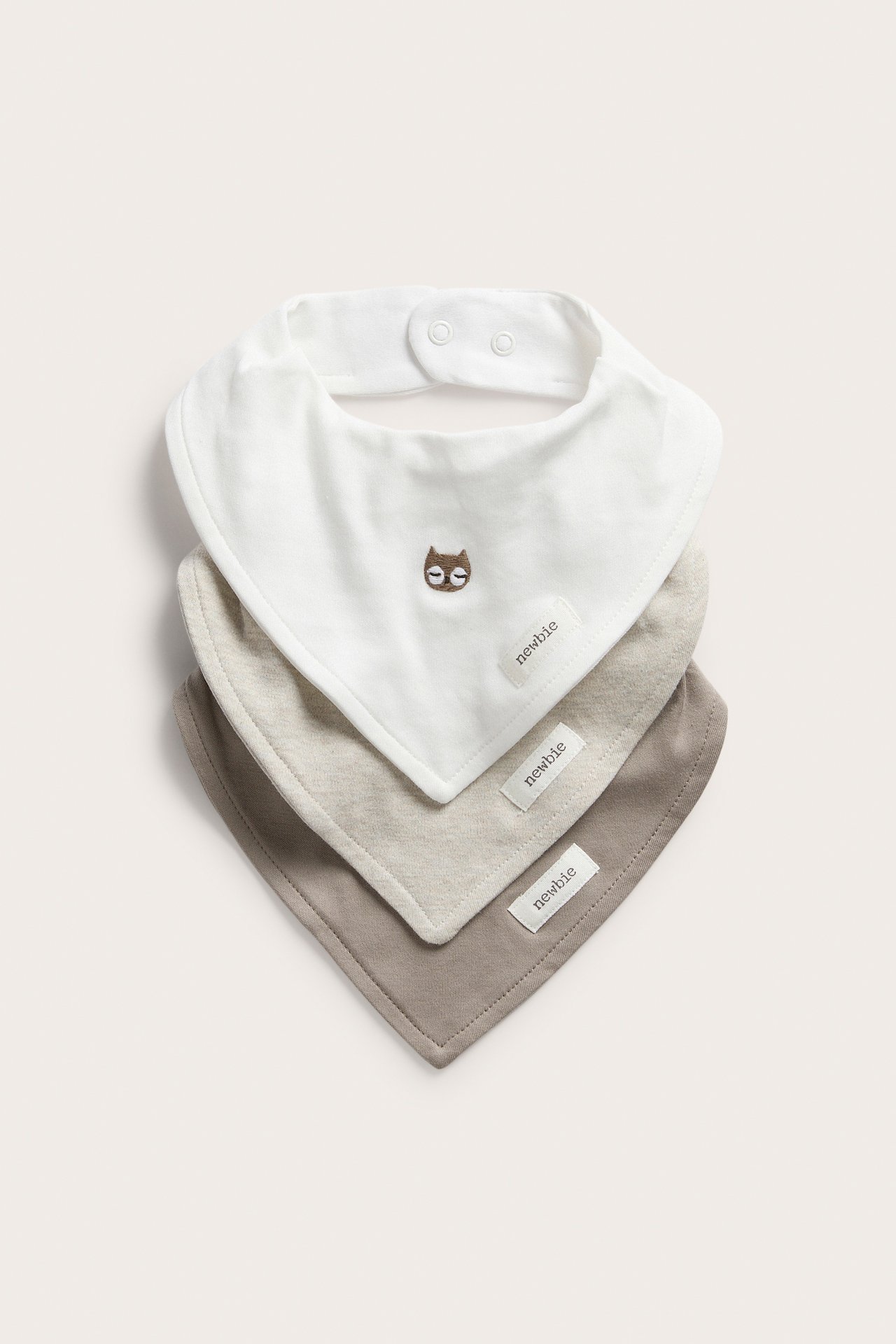 Front view of three stacked baby bibs: off-white with owl embroidery, light beige, and taupe.