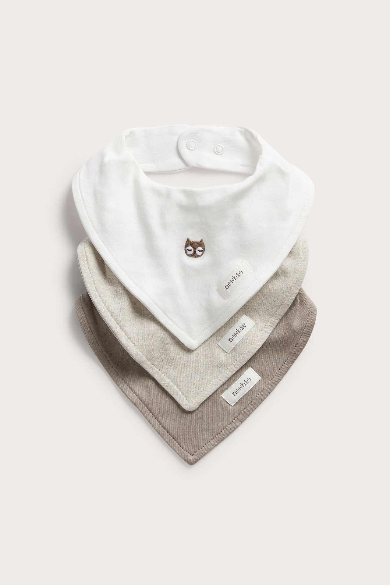 Vorderansicht von drei gestapelten Baby-Lätzchen: cremeweiß mit Eulenstickerei, hellbeige und taupe.