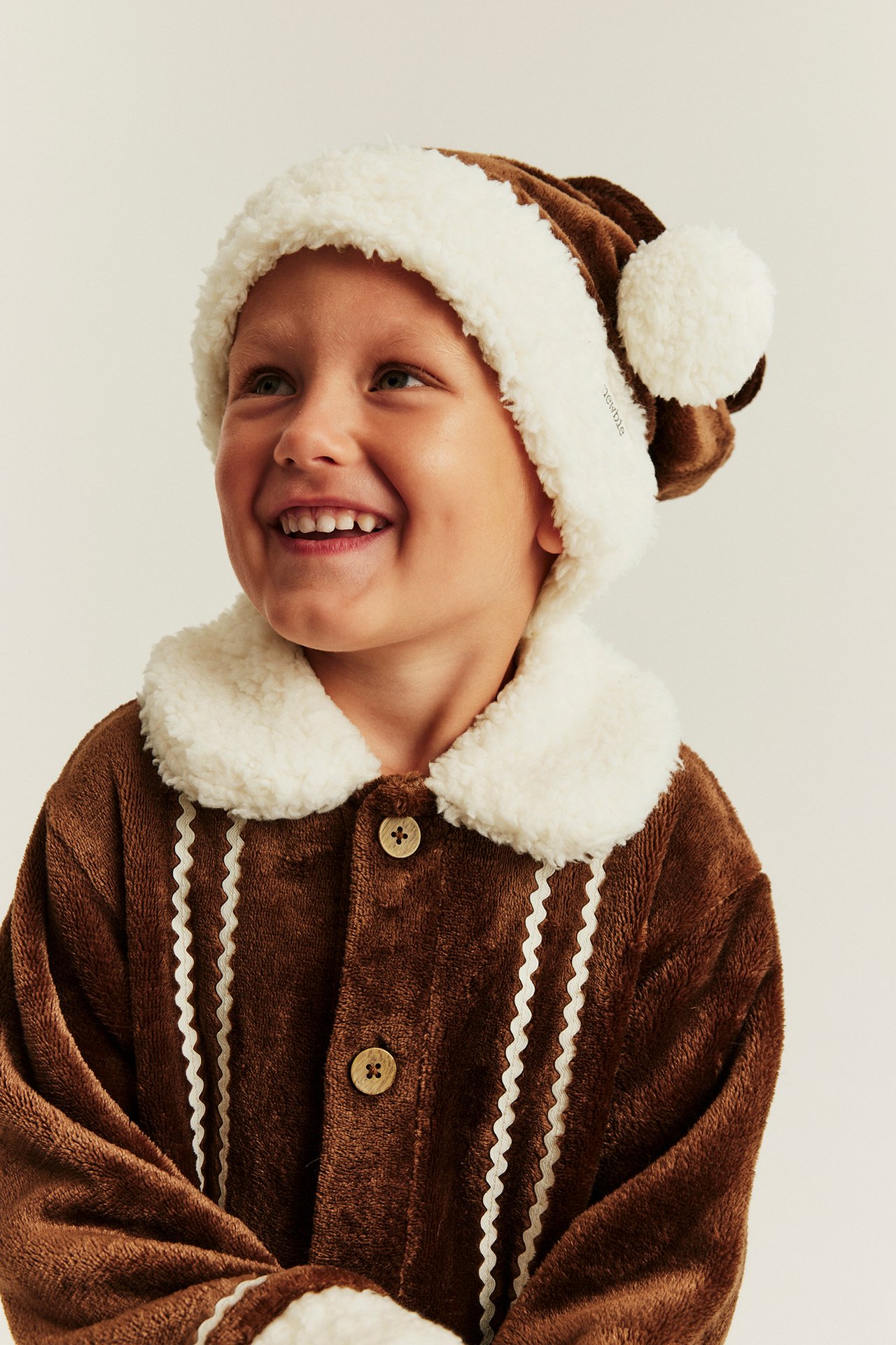 Vorderansicht Kind in braunem Plüsch-Lebkuchenmann-Pyjama-Set mit Knöpfen und Mütze.