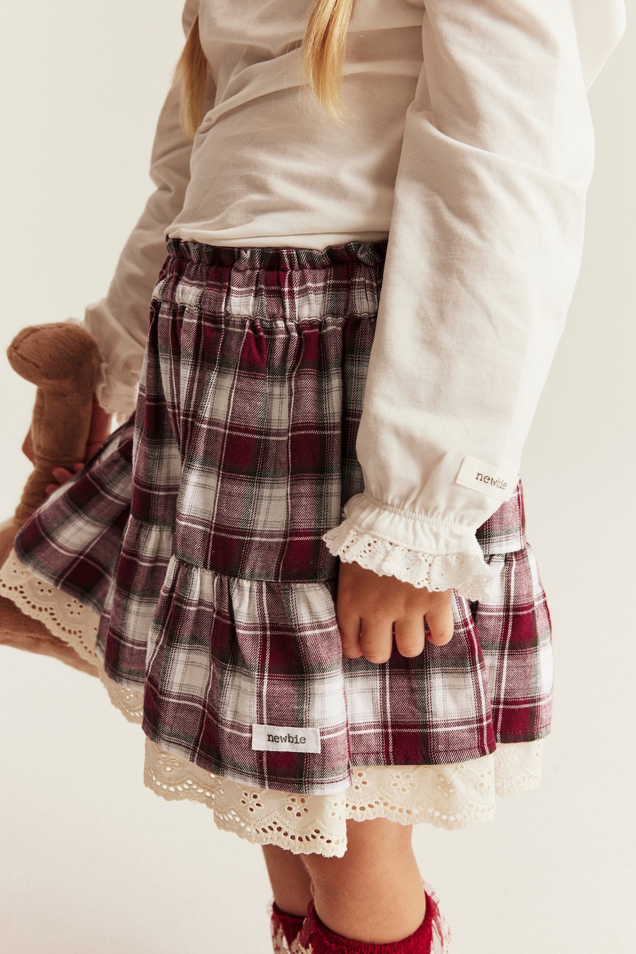 Kid in red plaid tiered skirt with white lace, white top, and a brown teddy bear.