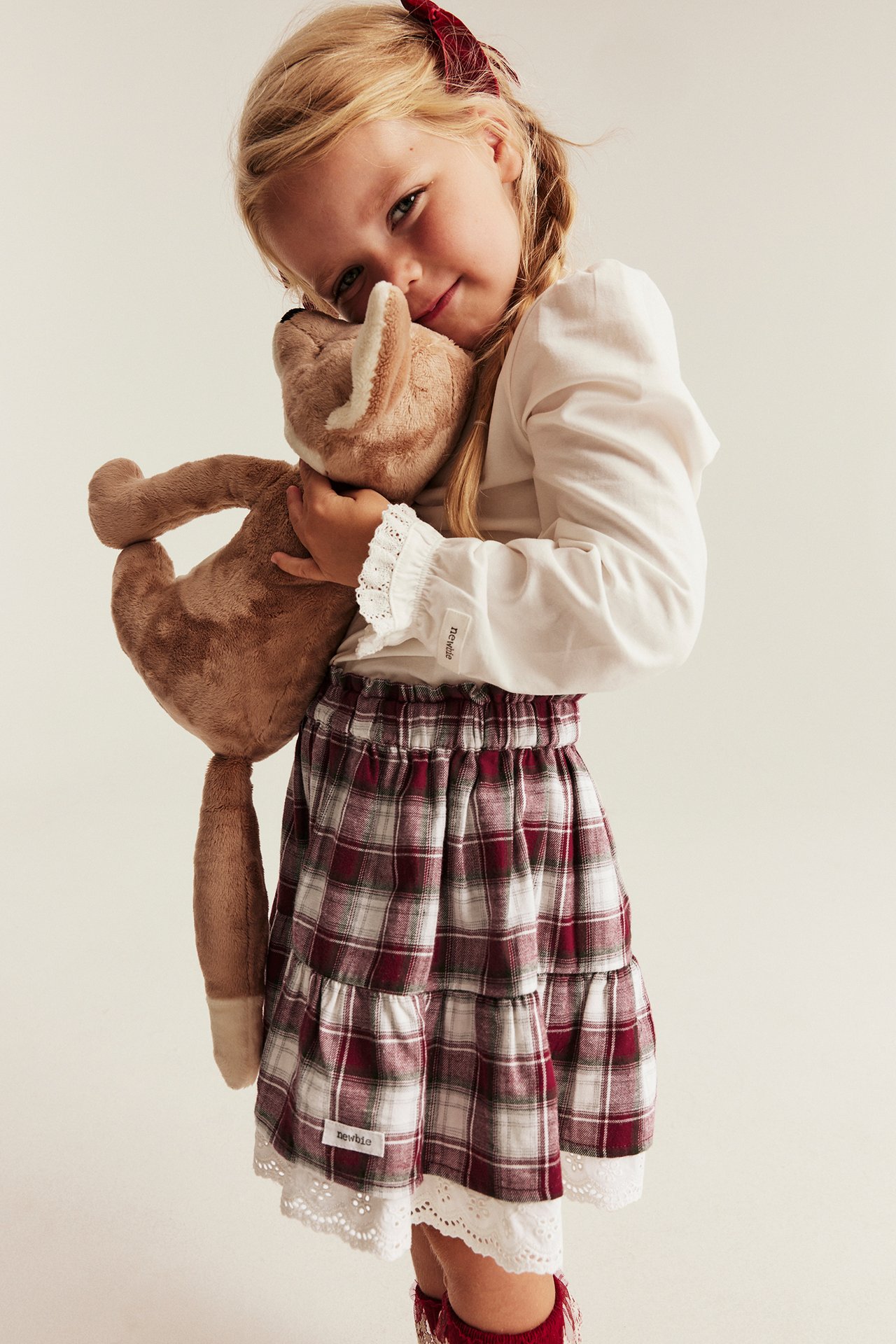 Front view: Kid's red plaid tiered skirt, lace trim, white blouse, red bow, holding a stuffed animal.