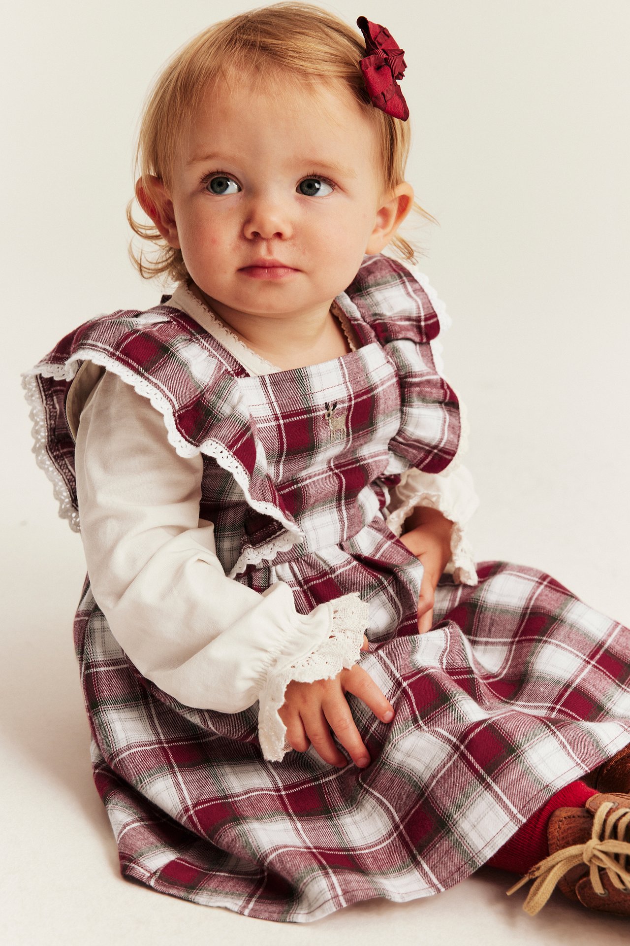 Front view of baby in red plaid pinafore dress over white long-sleeved top with lace cuffs.