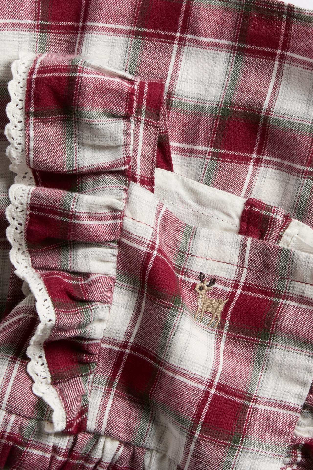 Close-up of a kid's red, white, and green plaid garment with ruffled straps and a deer embroidery.