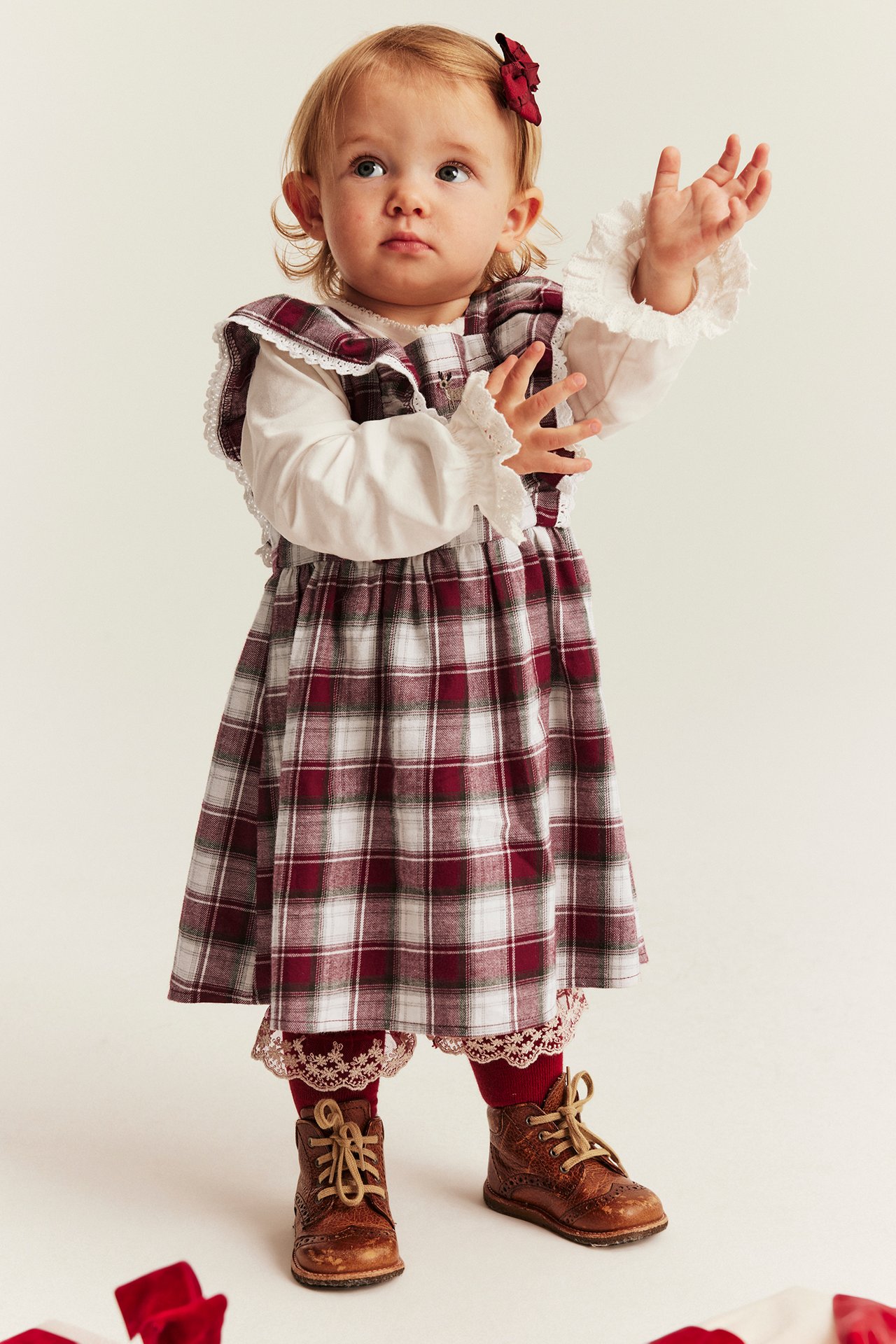 Front view: Baby in red plaid dress, white ruffled blouse, red lace tights, brown boots, and red bow.