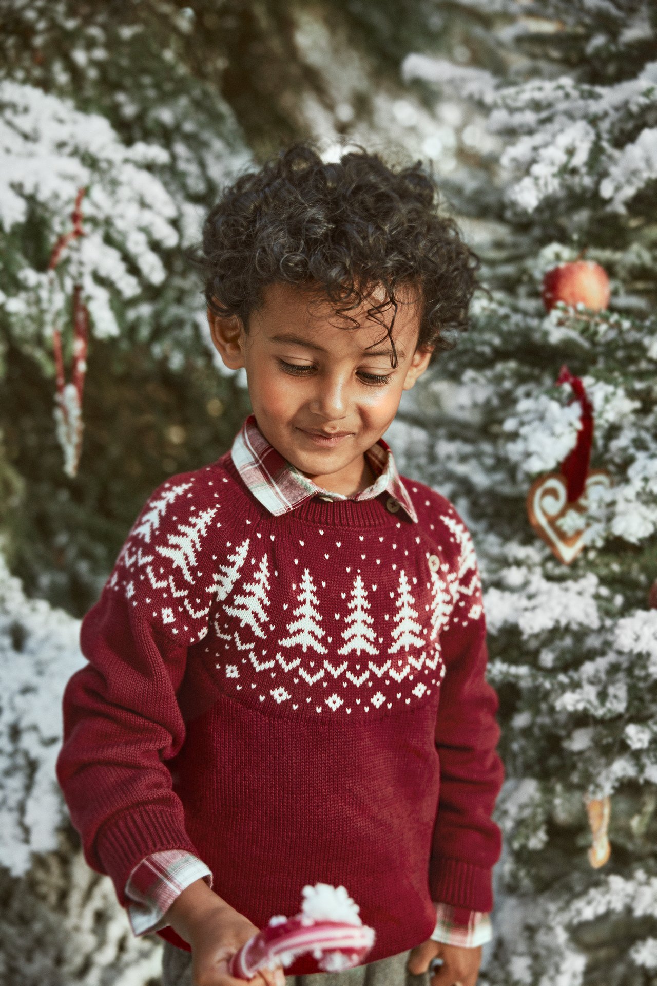 Vorderansicht: Kind in rotem Pullover mit Tannenbaummuster über kariertem Hemd, hält Zuckerstange.