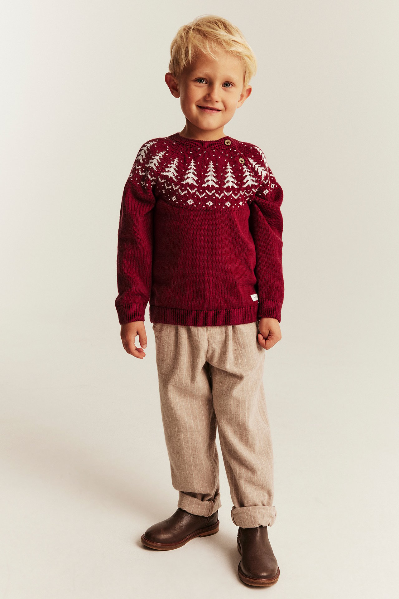 Front view of a kid wearing a red Christmas tree sweater, beige striped pants, and brown ankle boots.