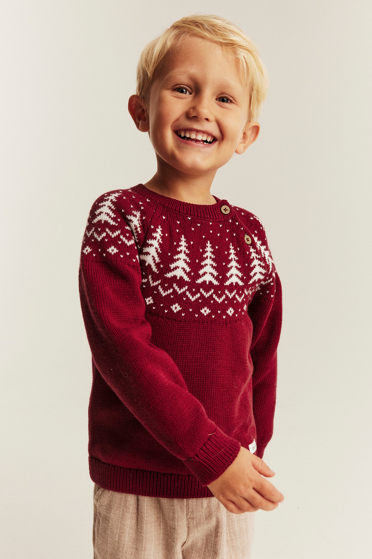Kid in red knit sweater with white fir trees, shoulder buttons, and beige striped pants.