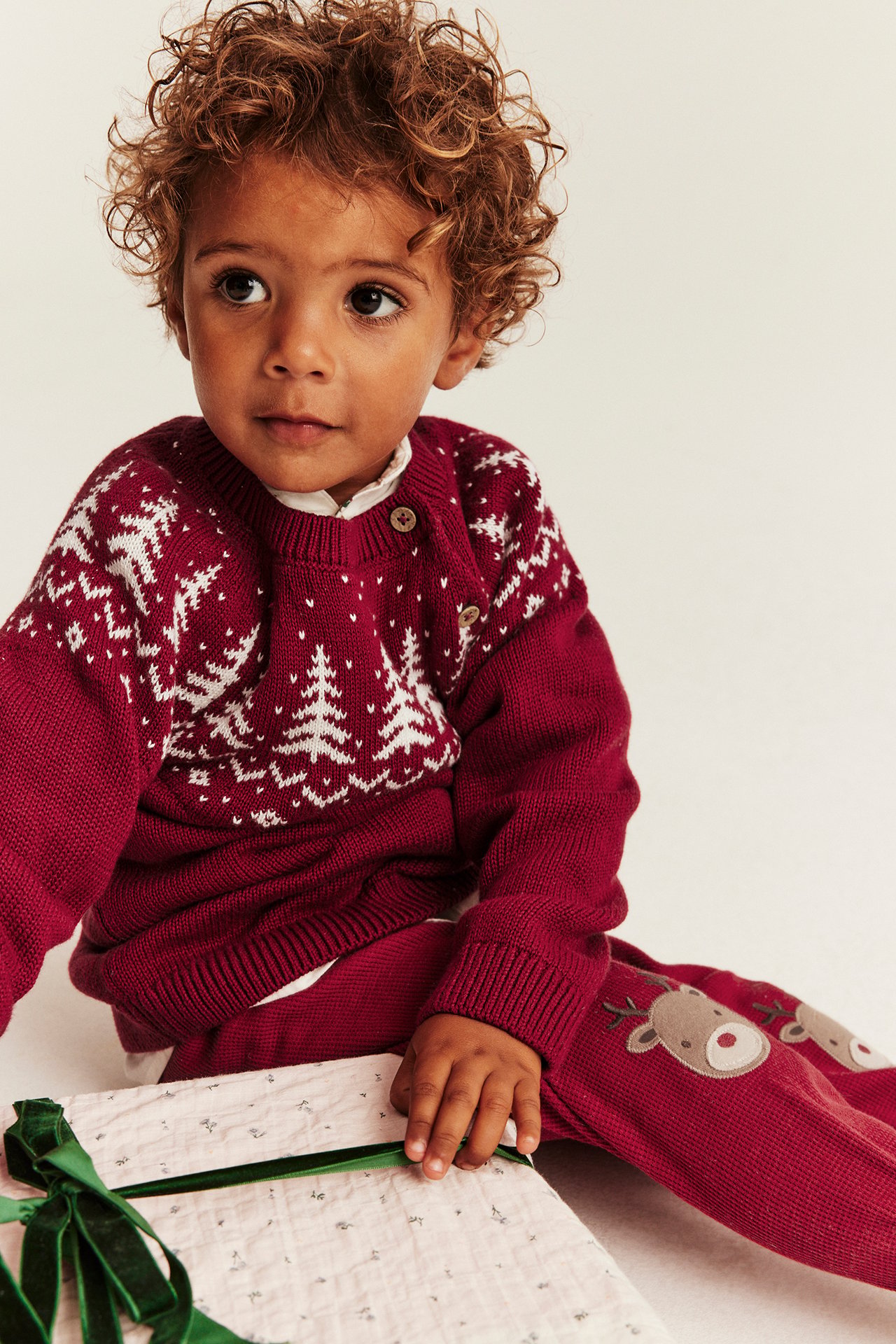 Vorderansicht: Kind in rotem Strickpullover mit weißem Wintermuster und passender Rentierhose, hält ein Geschenk.