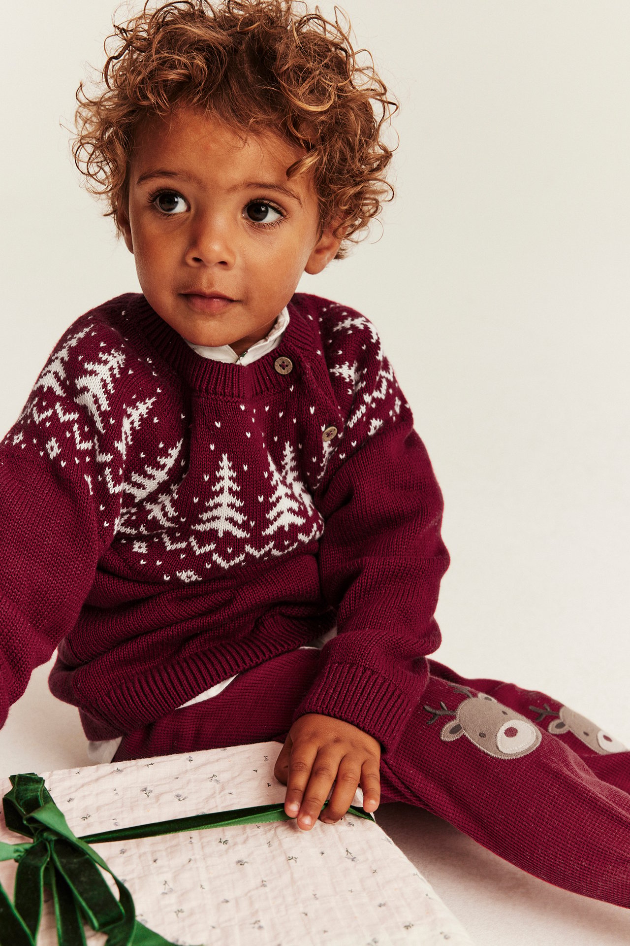 Vorderansicht eines Babys in weinrotem Strickpullover mit Tannenbaum-Muster und Rentier-Hose, hält ein Geschenk.