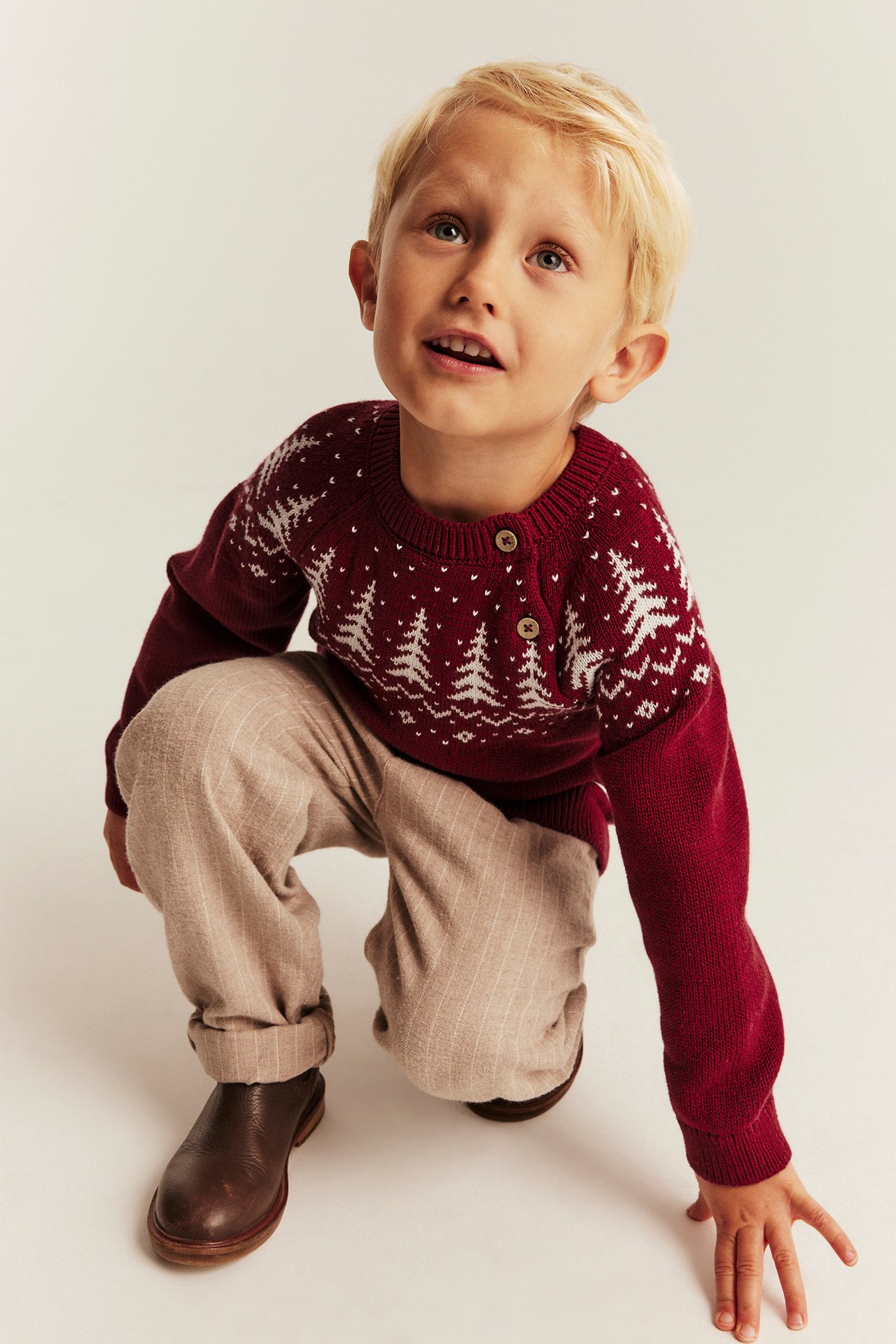 Vorderansicht, Kind in weinrotem Strickpullover mit weißen Tannen, beiger gestreifter Hose, braunen Stiefeln.
