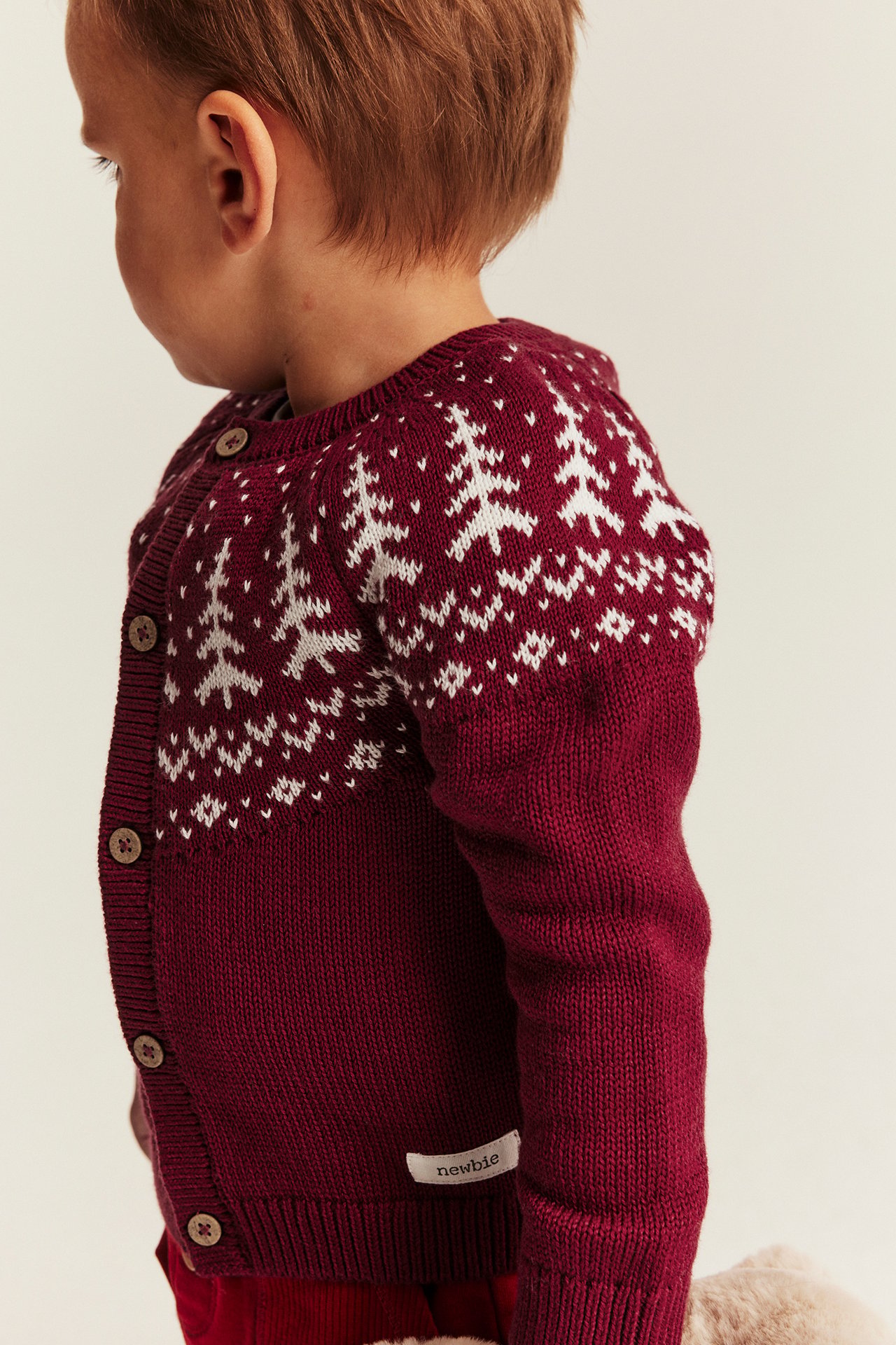 Seitenansicht, Nahaufnahme eines Babys in roter Strickjacke mit weißem Fair Isle-Muster und braunen Knöpfen.