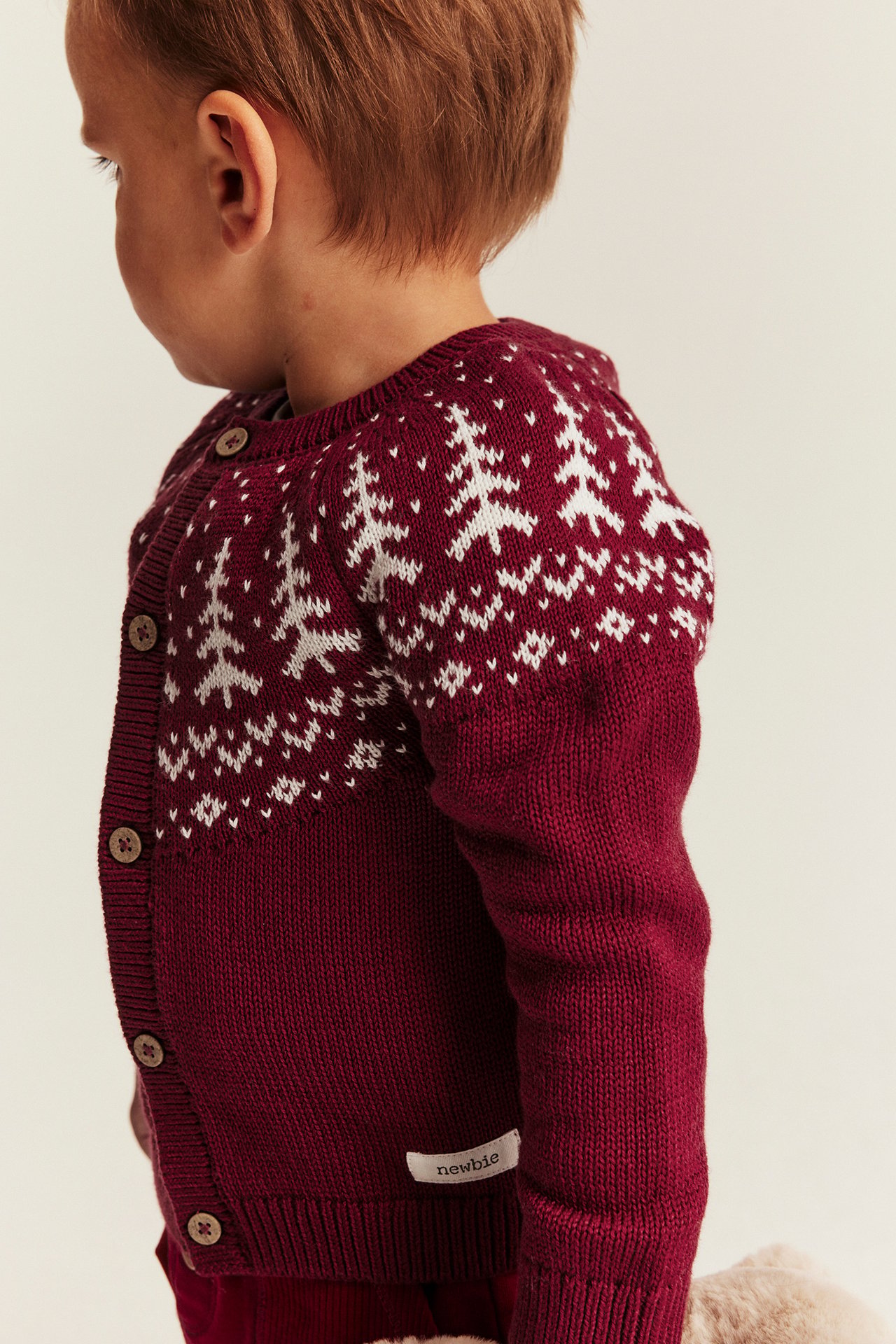 Seitenansicht eines Babys in dunkelroter Strickjacke mit weißem Fair-Isle-Tannenbaum-Muster.