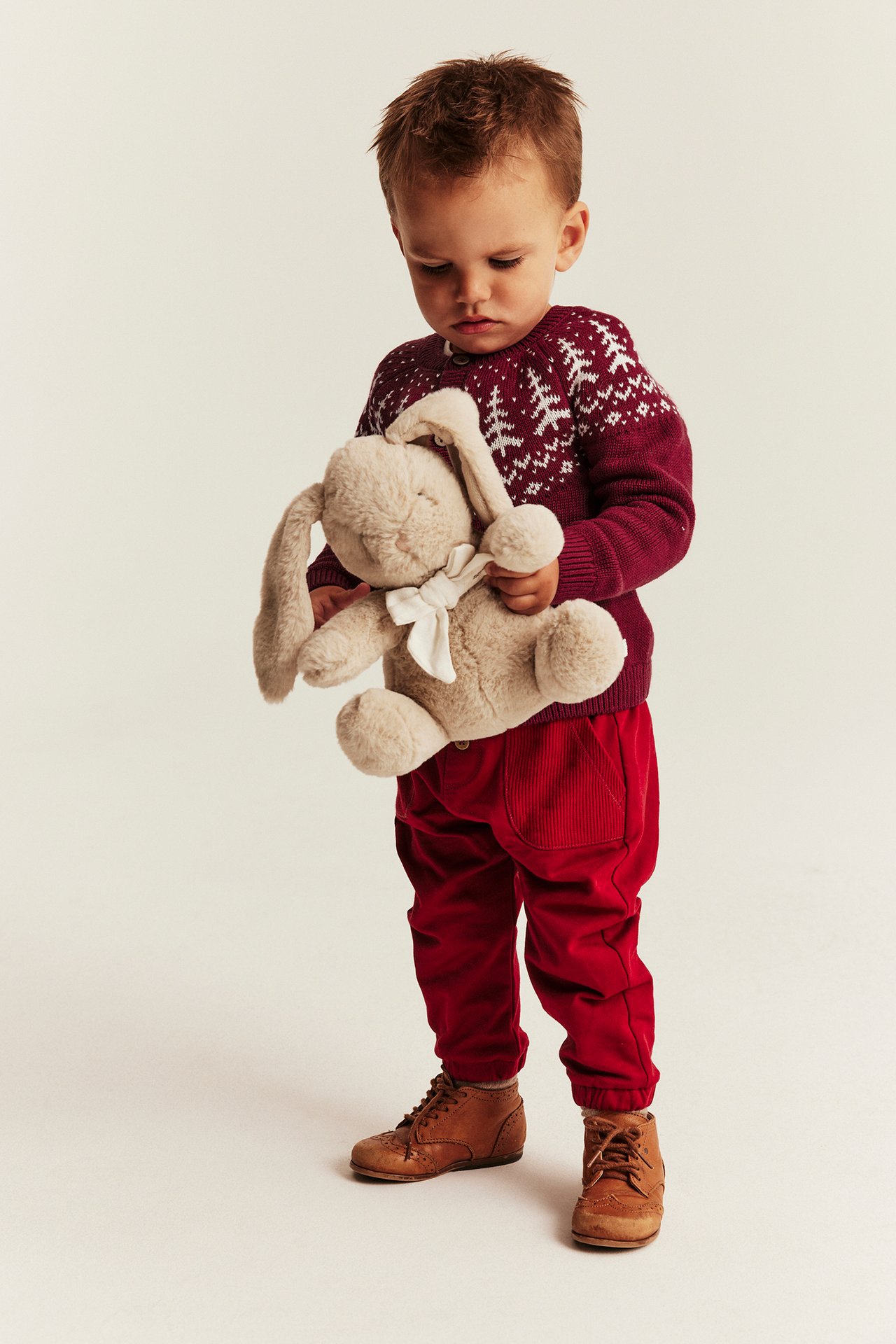 Kind trägt roten Fair-Isle-Pullover, rote Cordhose, braune Schnürstiefel, hält Plüschhasen.