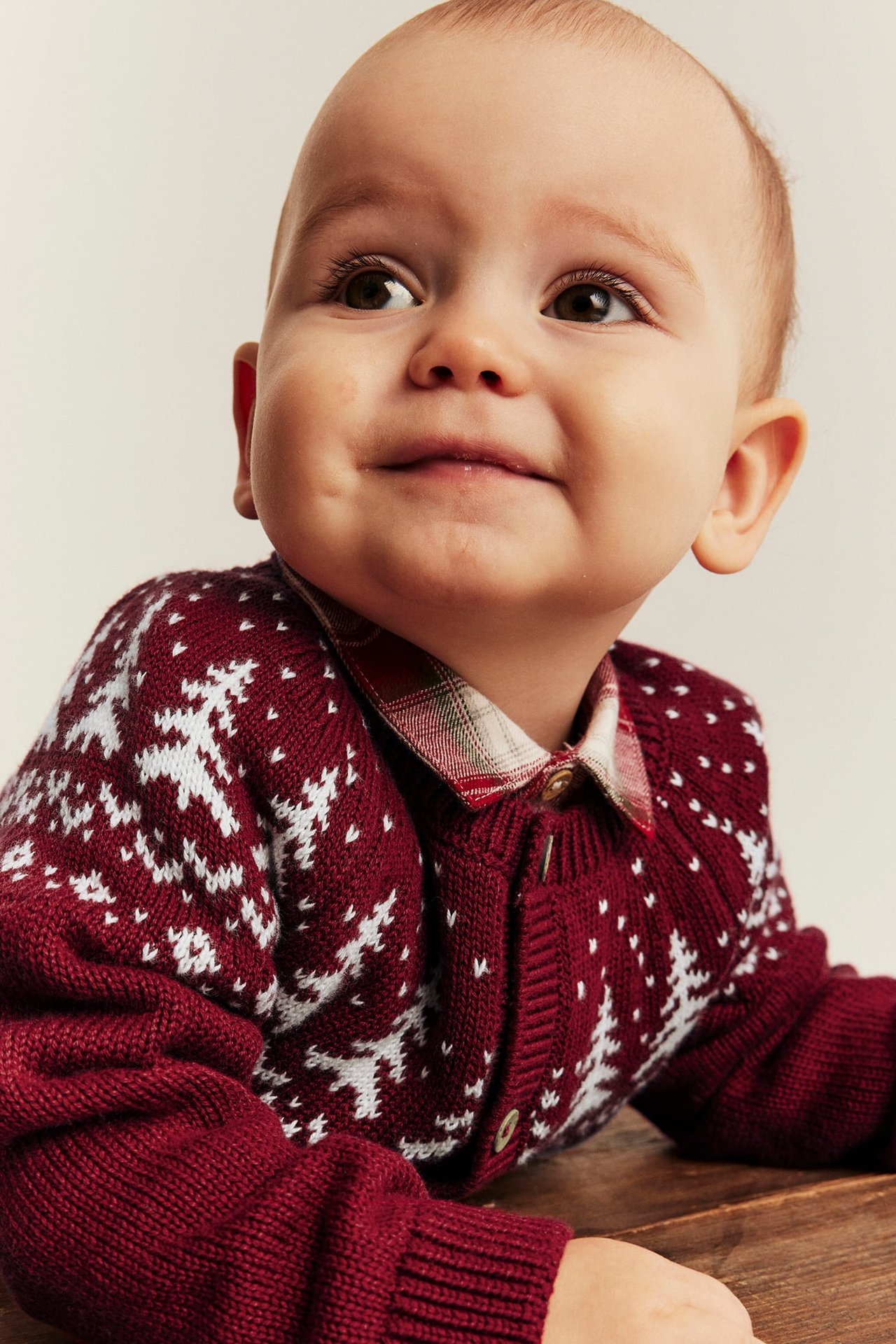 Close-up of a baby wearing a maroon knit cardigan with white snowflake pattern over a plaid shirt.