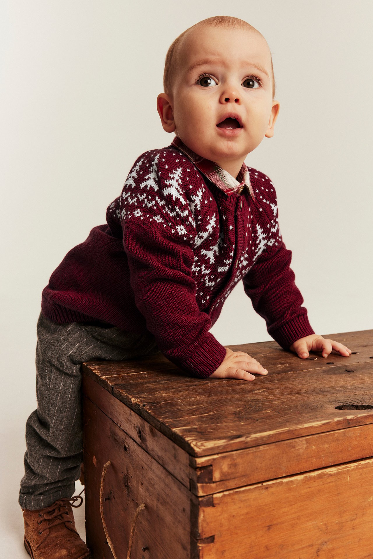 Front view of a baby wearing a maroon patterned cardigan, plaid shirt, gray pants, and brown shoes.