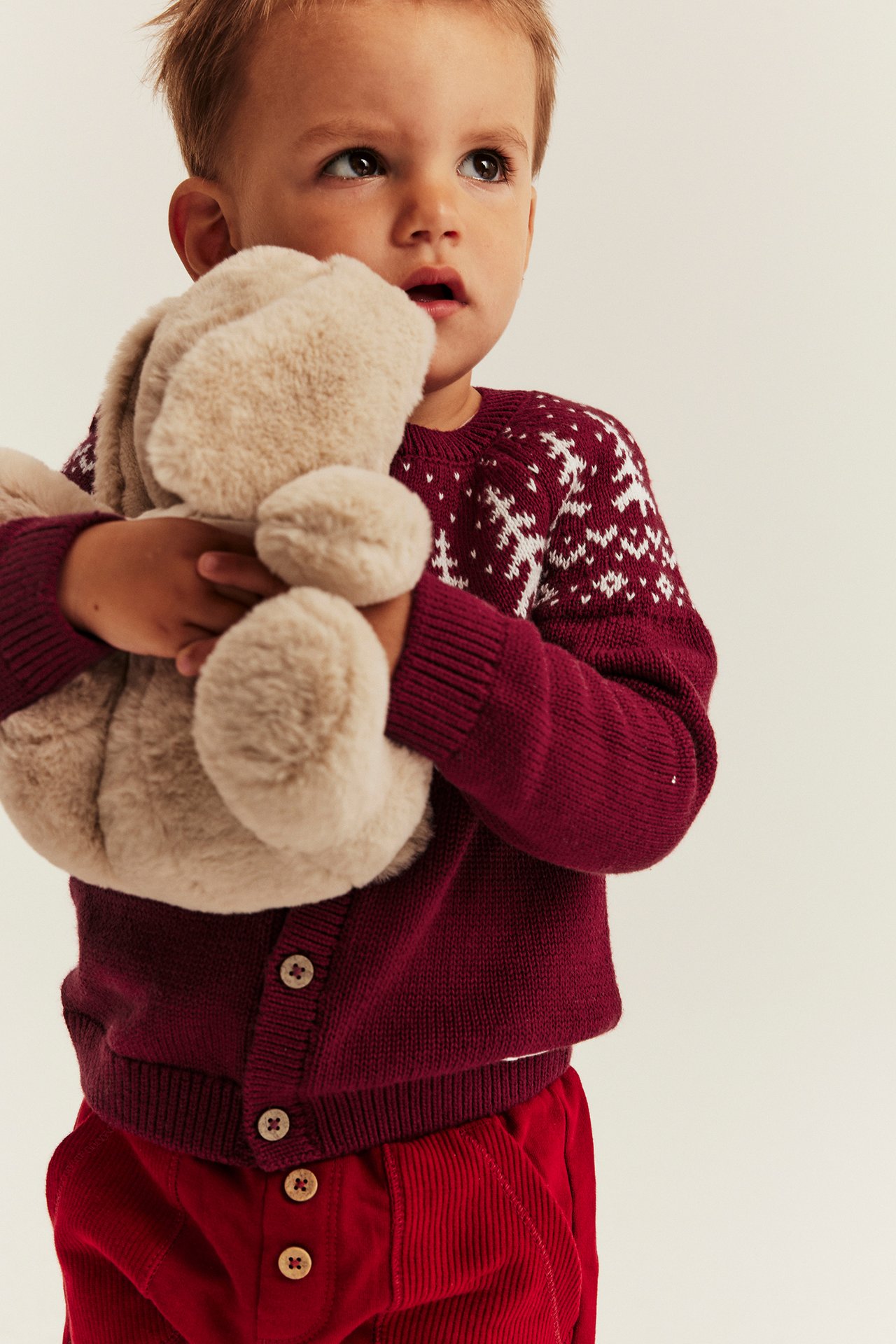 Frontansicht, Kind in burgunderroter Strickjacke, Schneeflockenpullover, roten Cordhosen, hält beiges Häschen.