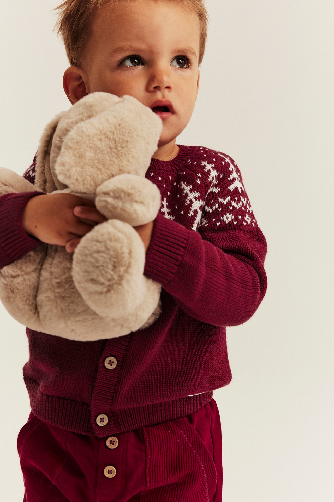Baby in burgunderroter Strickjacke mit Schneeflockenmuster und Cordhose, hält beiges Plüschtier.