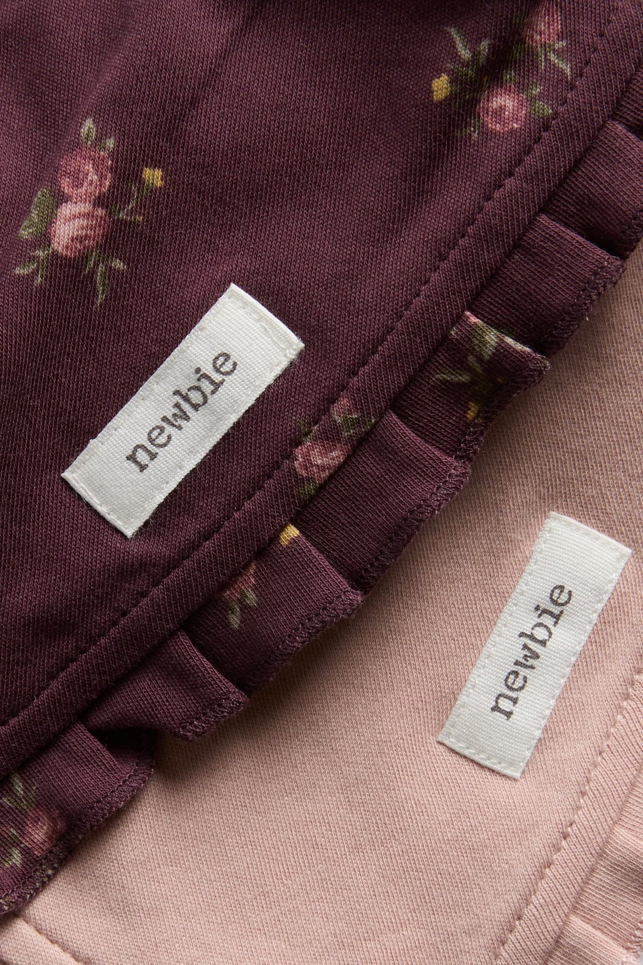 Nahaufnahme eines bordeauxroten Babykleidungsstücks mit Blumenmuster und Rüschen und eines rosa einfarbigen Babykleidungsstücks.