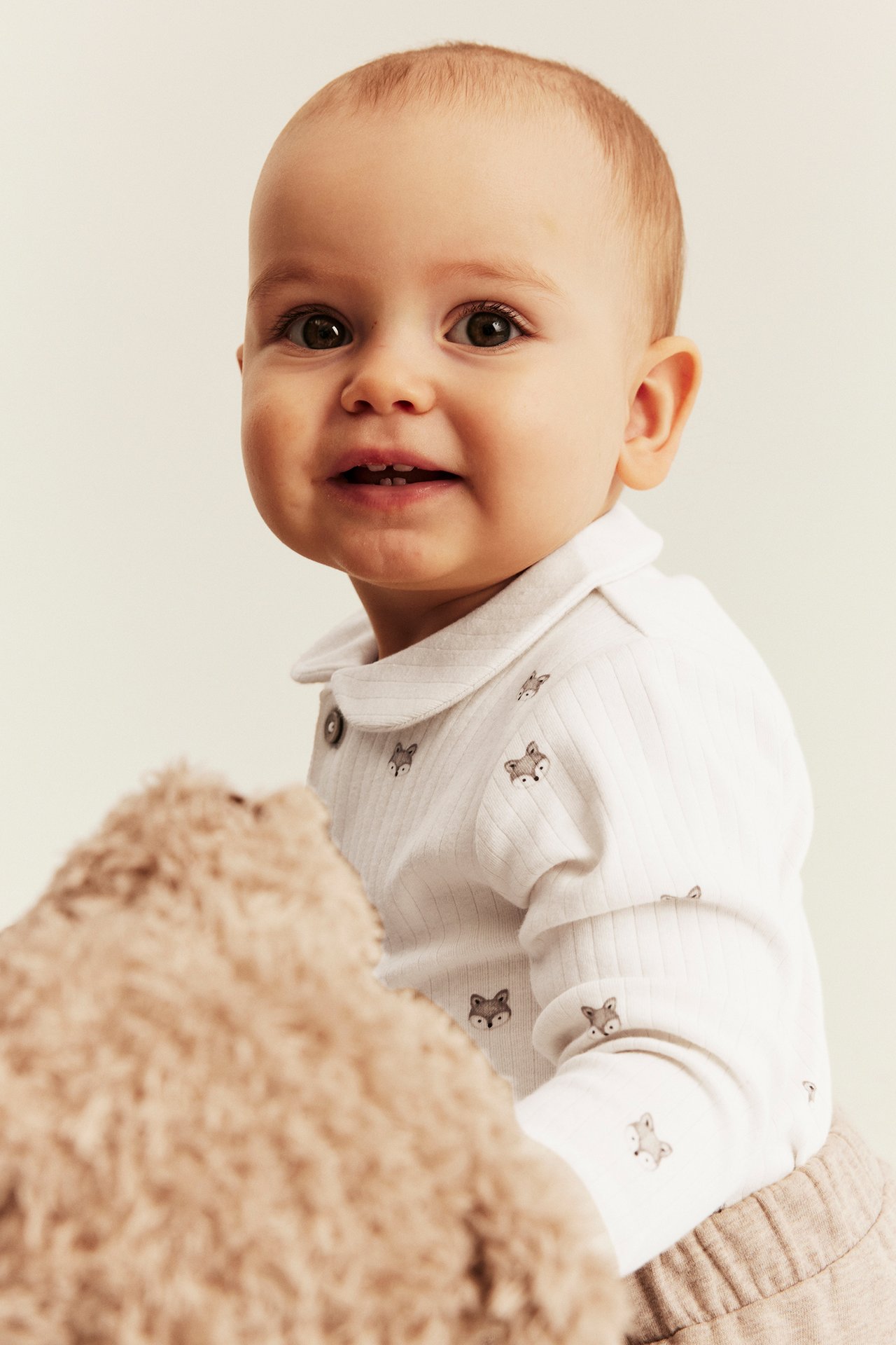 Vorderansicht: Baby in weißem geripptem Fuchsdruck-Langarmshirt und hellbrauner Hose, hält Kuscheltier.