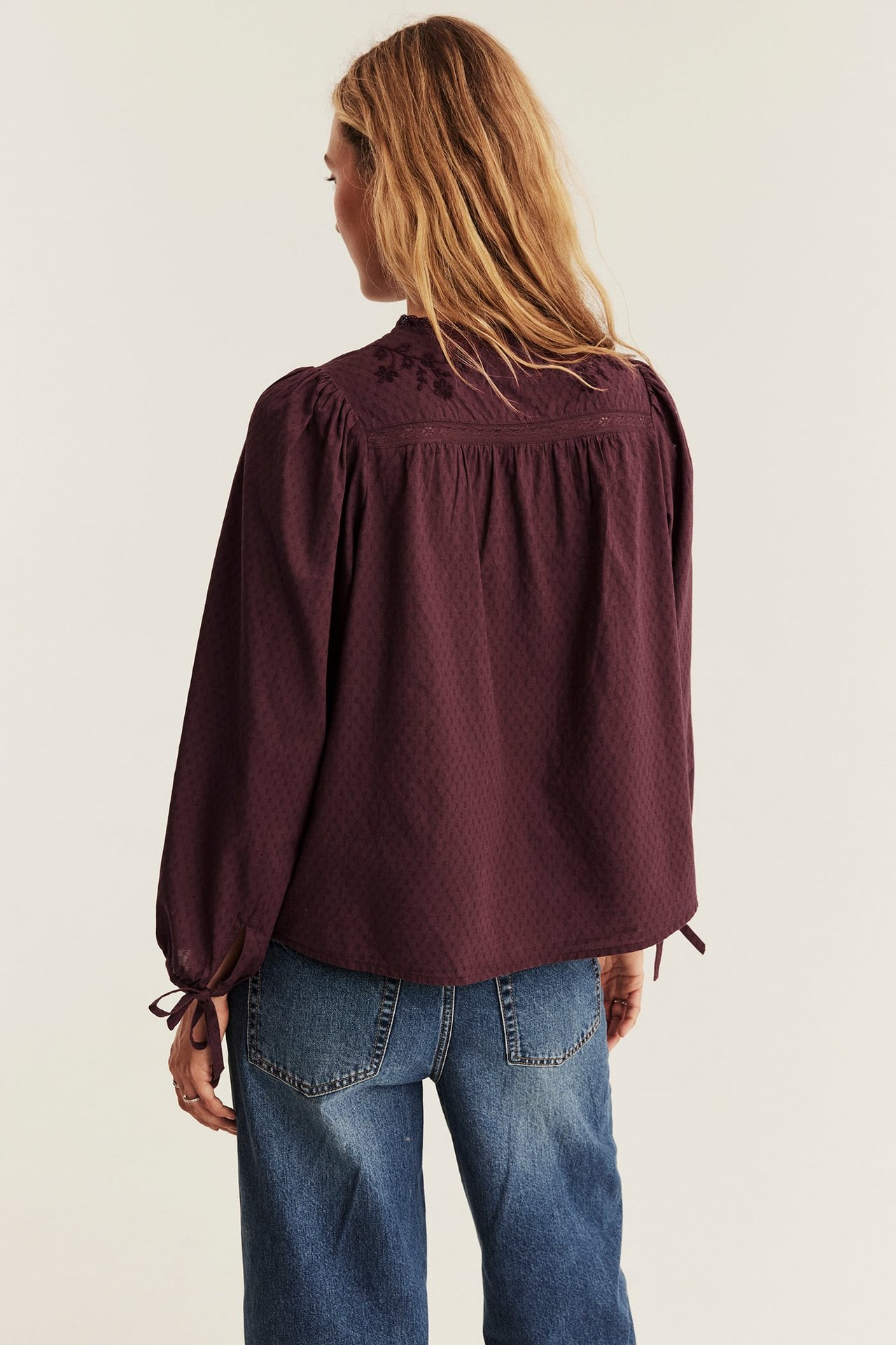 Back view of a woman wearing a maroon long-sleeve dotted embroidered blouse and blue denim jeans.