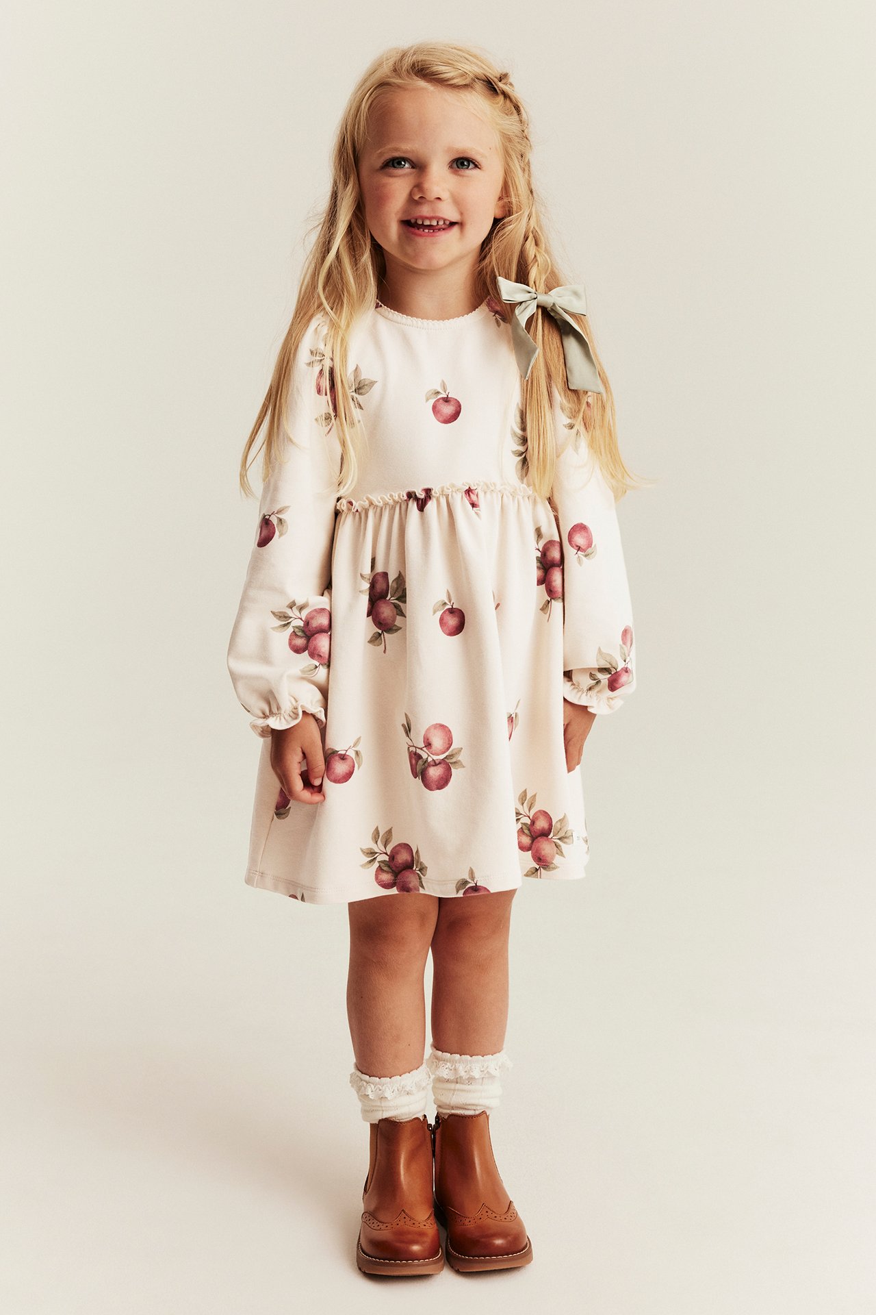 Front view of a kid in a cream long-sleeved dress with apple print, white ruffled socks, and brown boots.