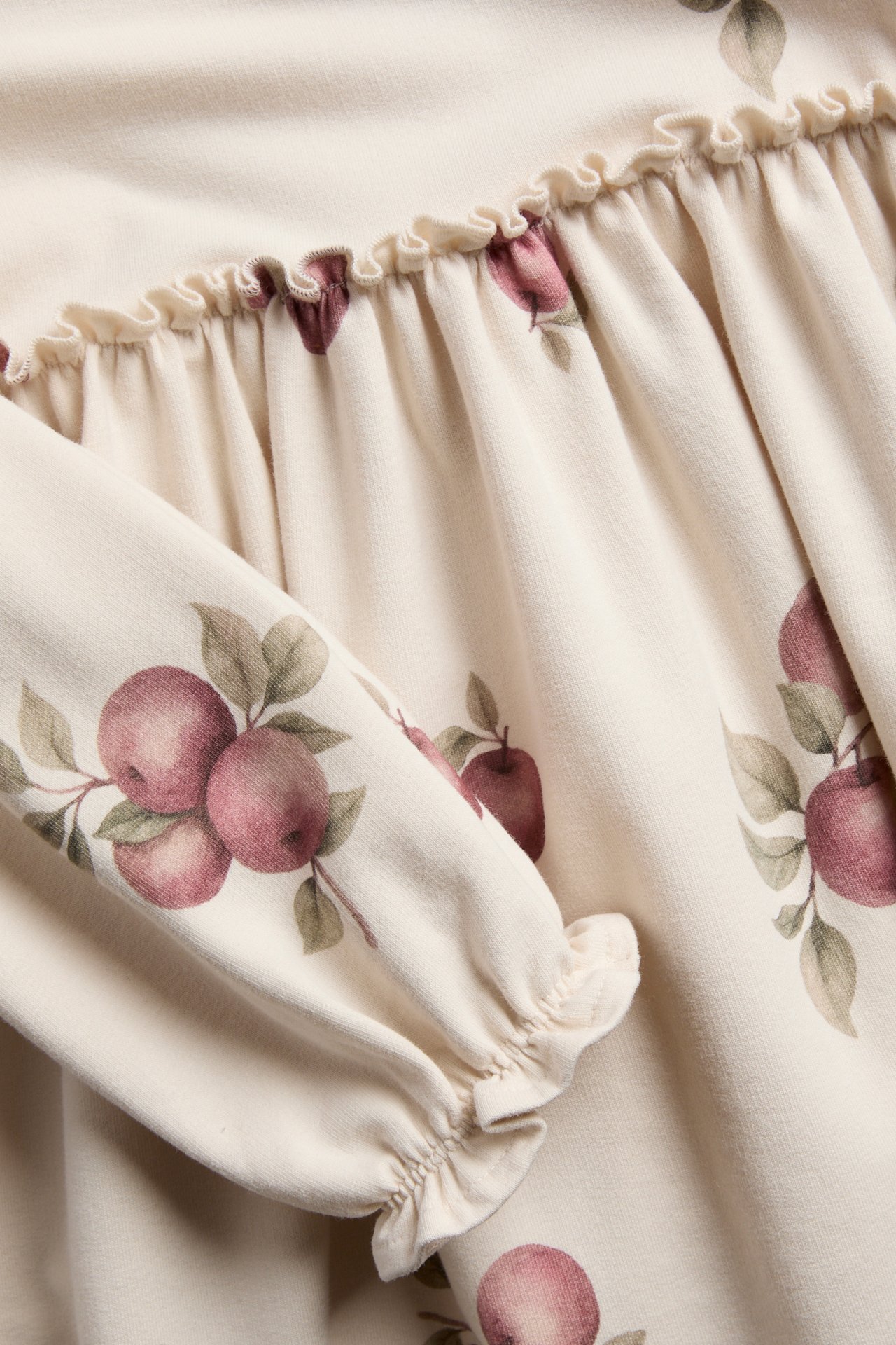 Nahaufnahme eines cremefarbenen Baby-Langarmshirts mit rotem Apfeldruck, Rüschenpasse und Rüschenbündchen.