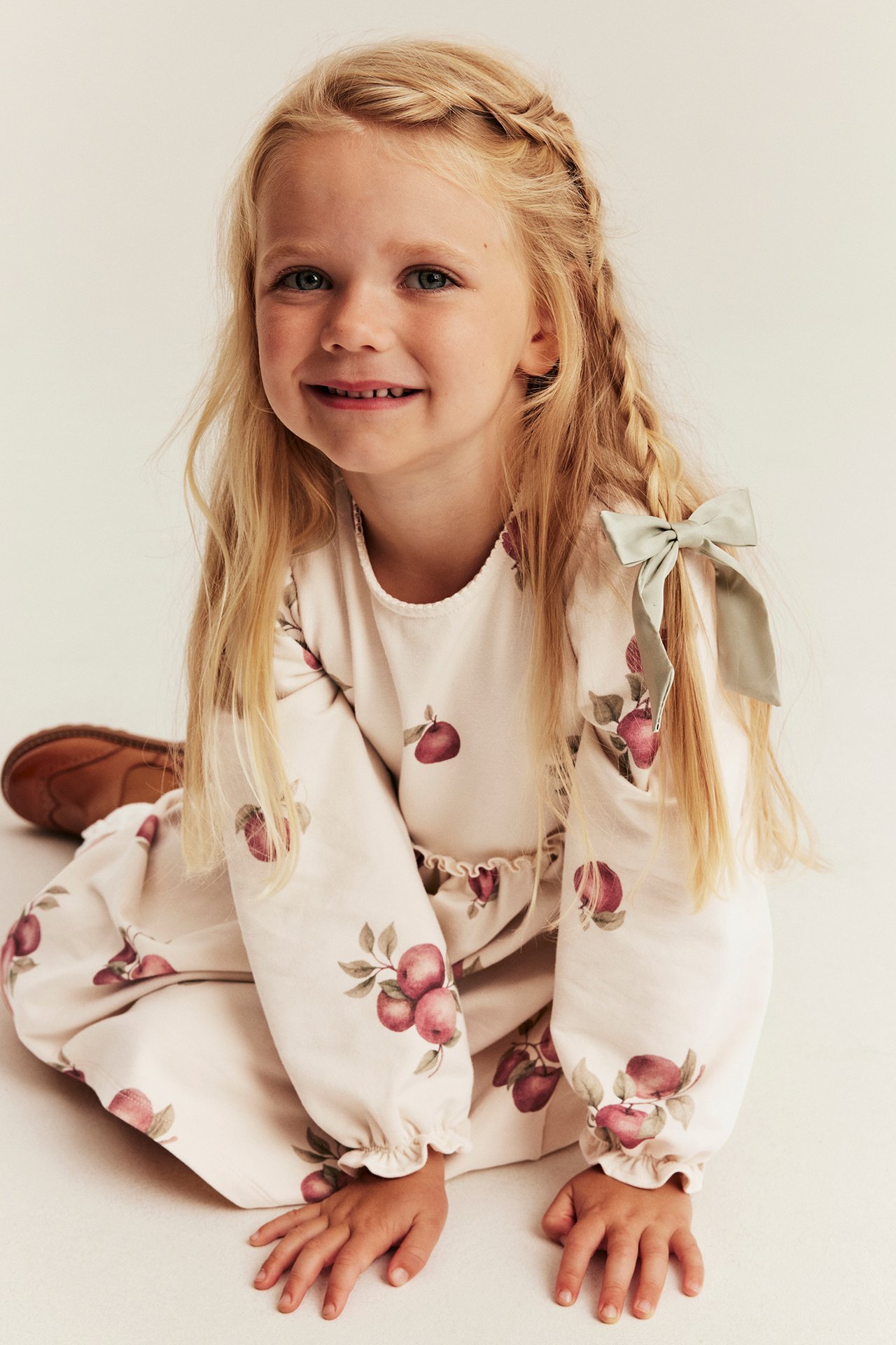 Front view of kid in cream long-sleeve dress with red apple print, light green hair bow, and brown shoes.