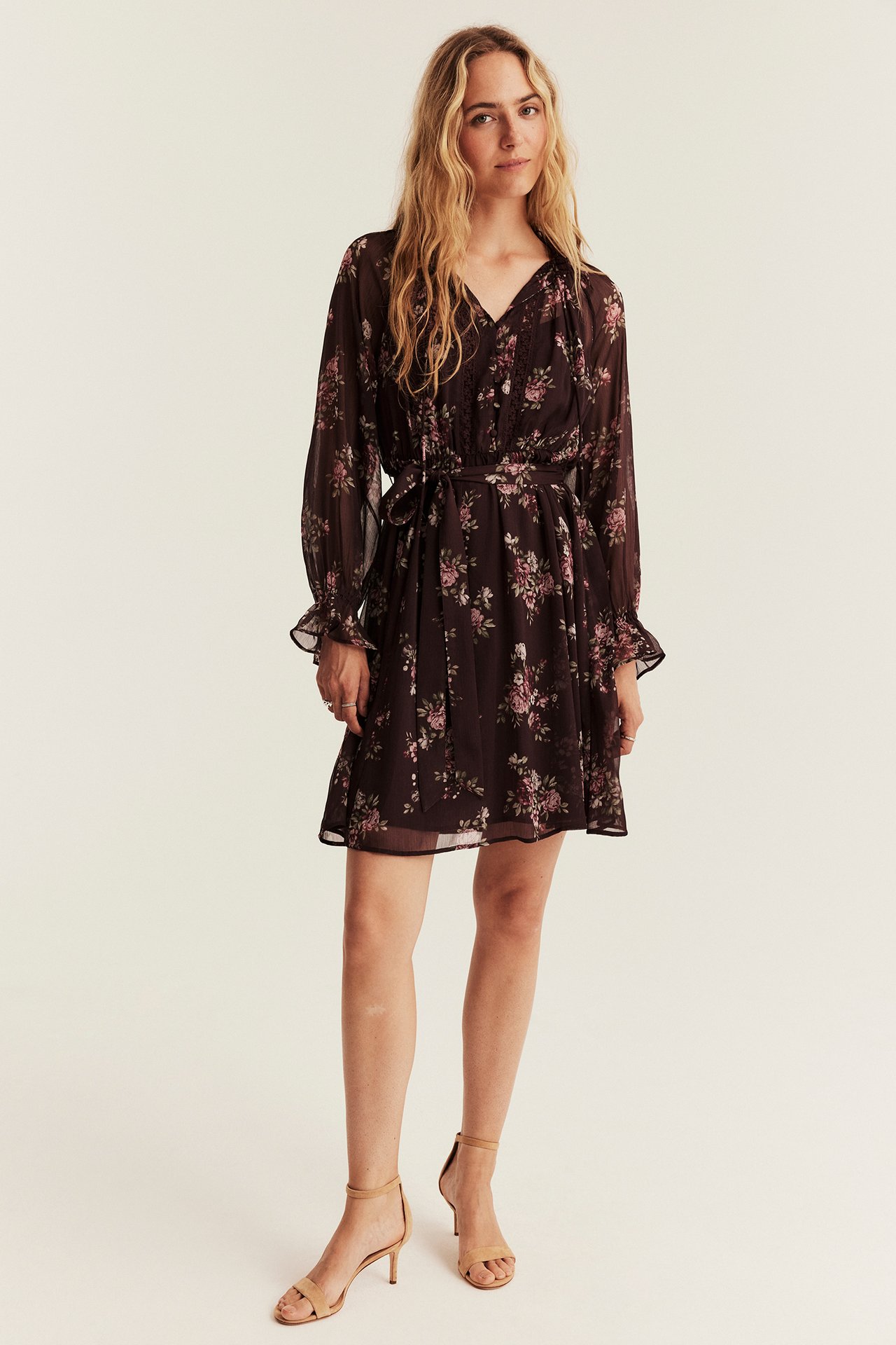 Front view: female model in a dark brown floral mini dress with tie waist and beige heels.