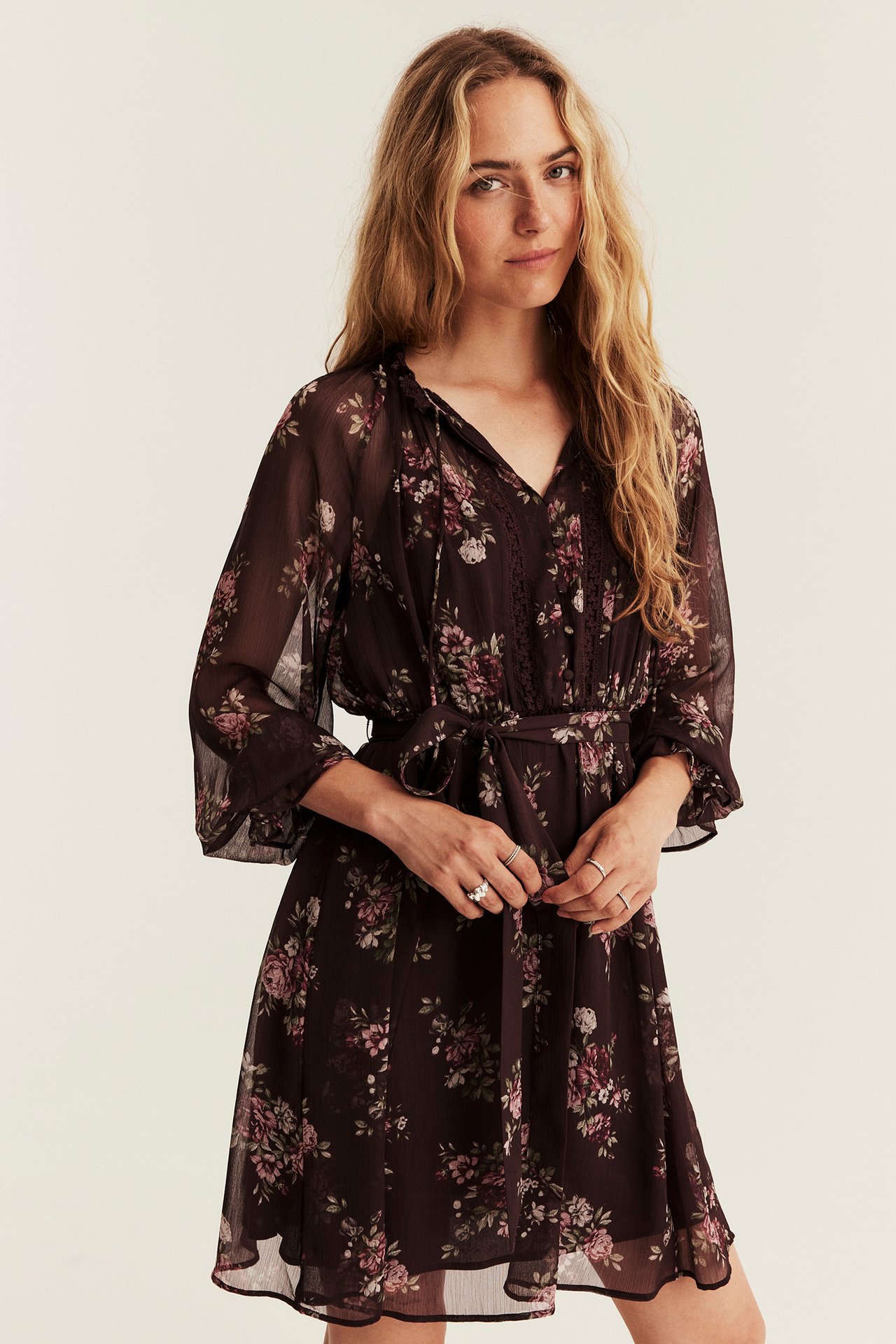 Front view of a female model in a dark brown floral long-sleeved dress with a V-neck and rings.