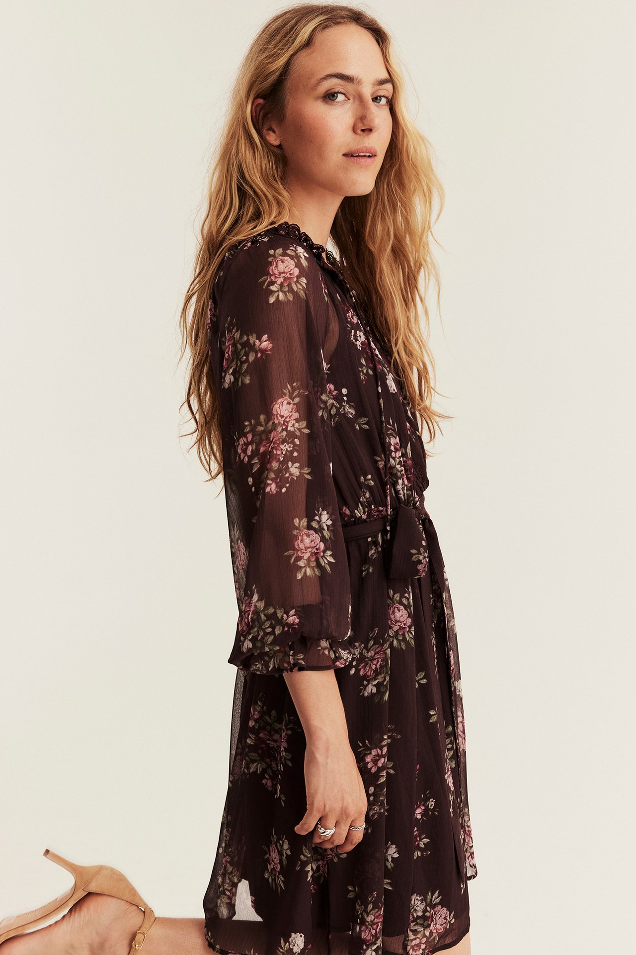 Side view of a female model in a dark floral sheer dress with tie waist and nude heels.