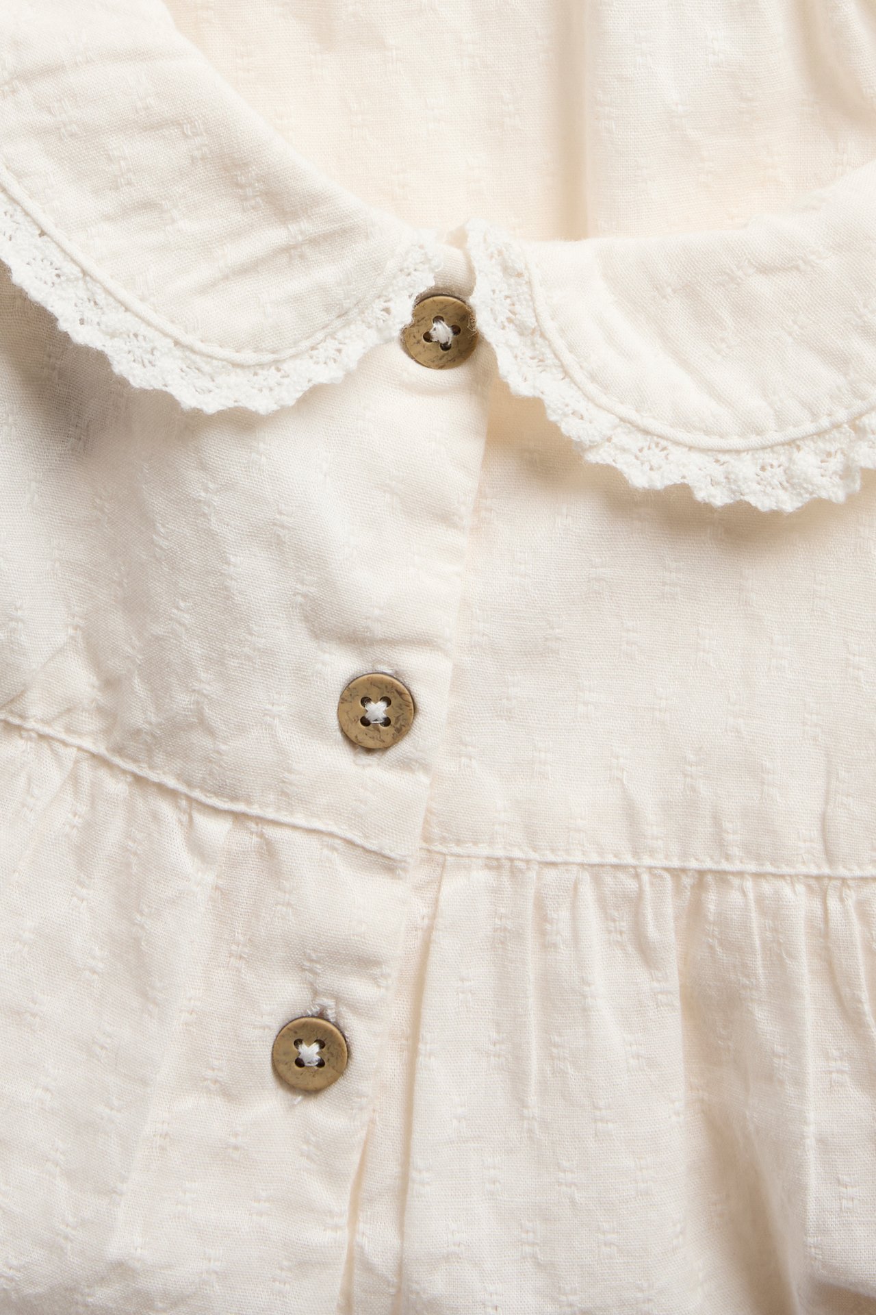 Close-up of a baby's cream textured dress with a lace-trimmed collar and brown buttons.