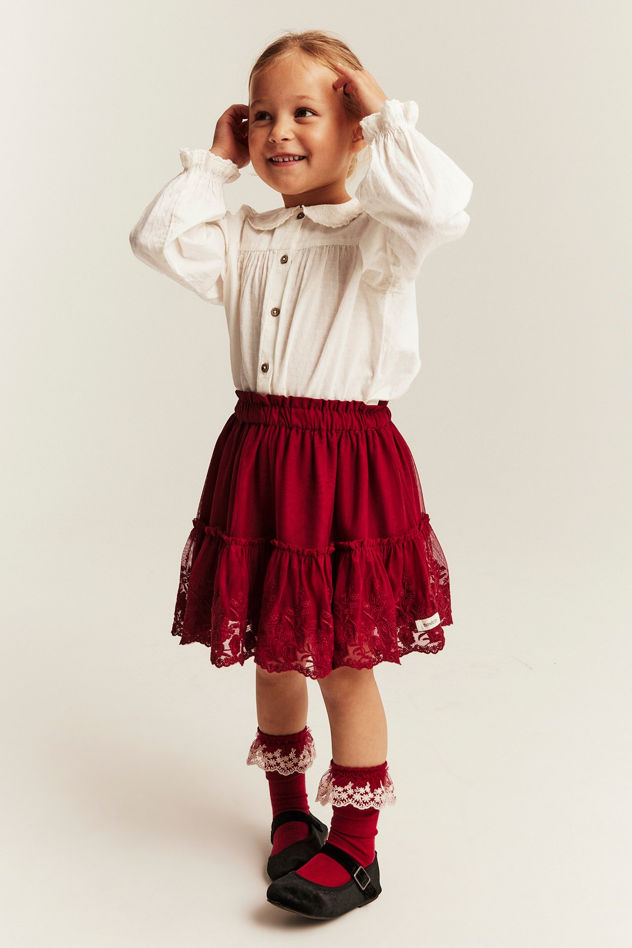 Front view of kid in red lace-tiered skirt, white long-sleeve blouse, red lace socks, black shoes.