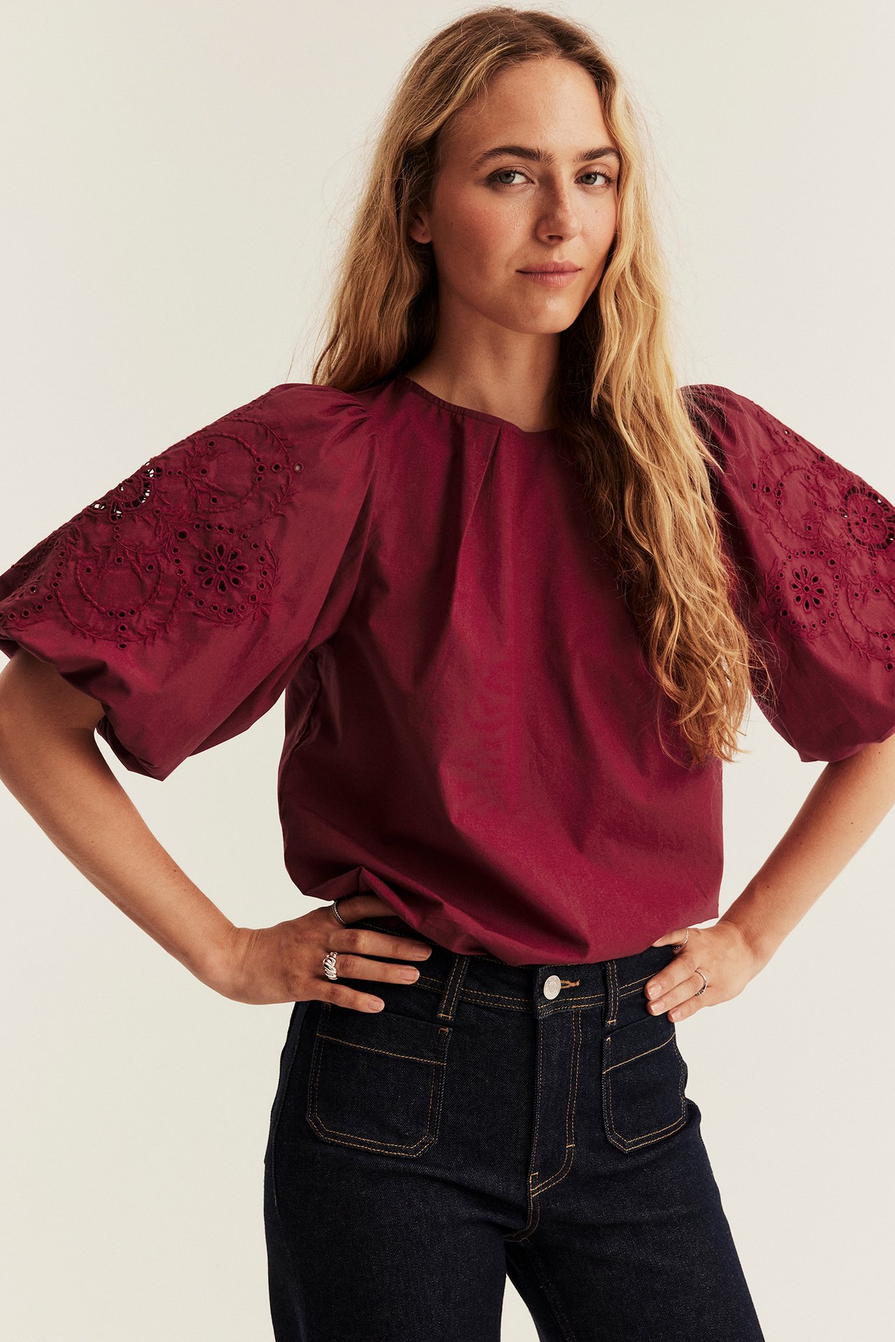 Vorderansicht einer Frau in einer weinroten, bestickten Puffärmelbluse und dunkelblauen Jeans.