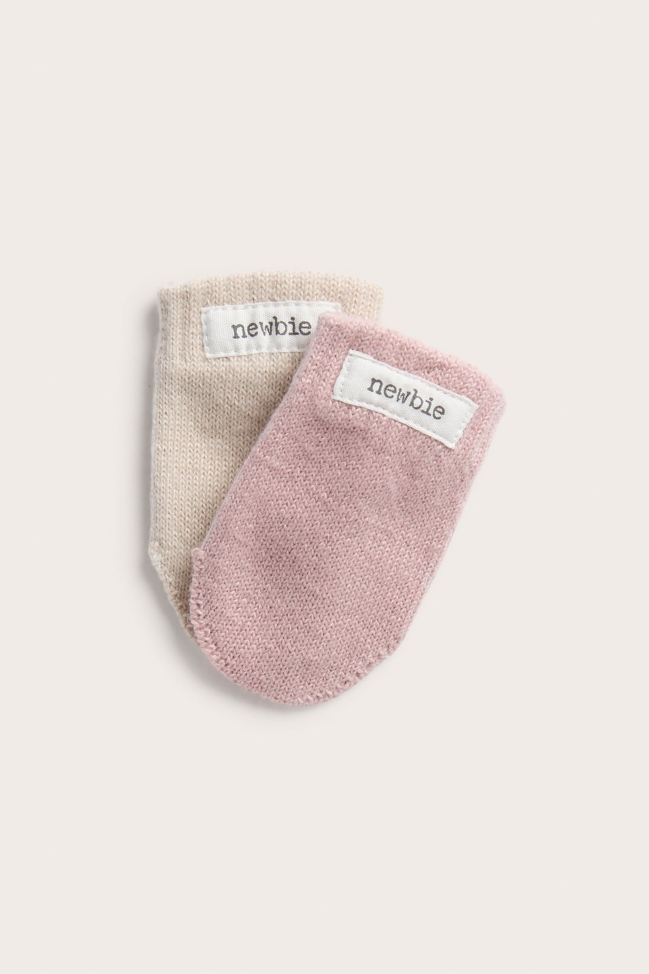 Close-up of two baby knitted mittens, one pink and one beige, with 'newbie' labels.