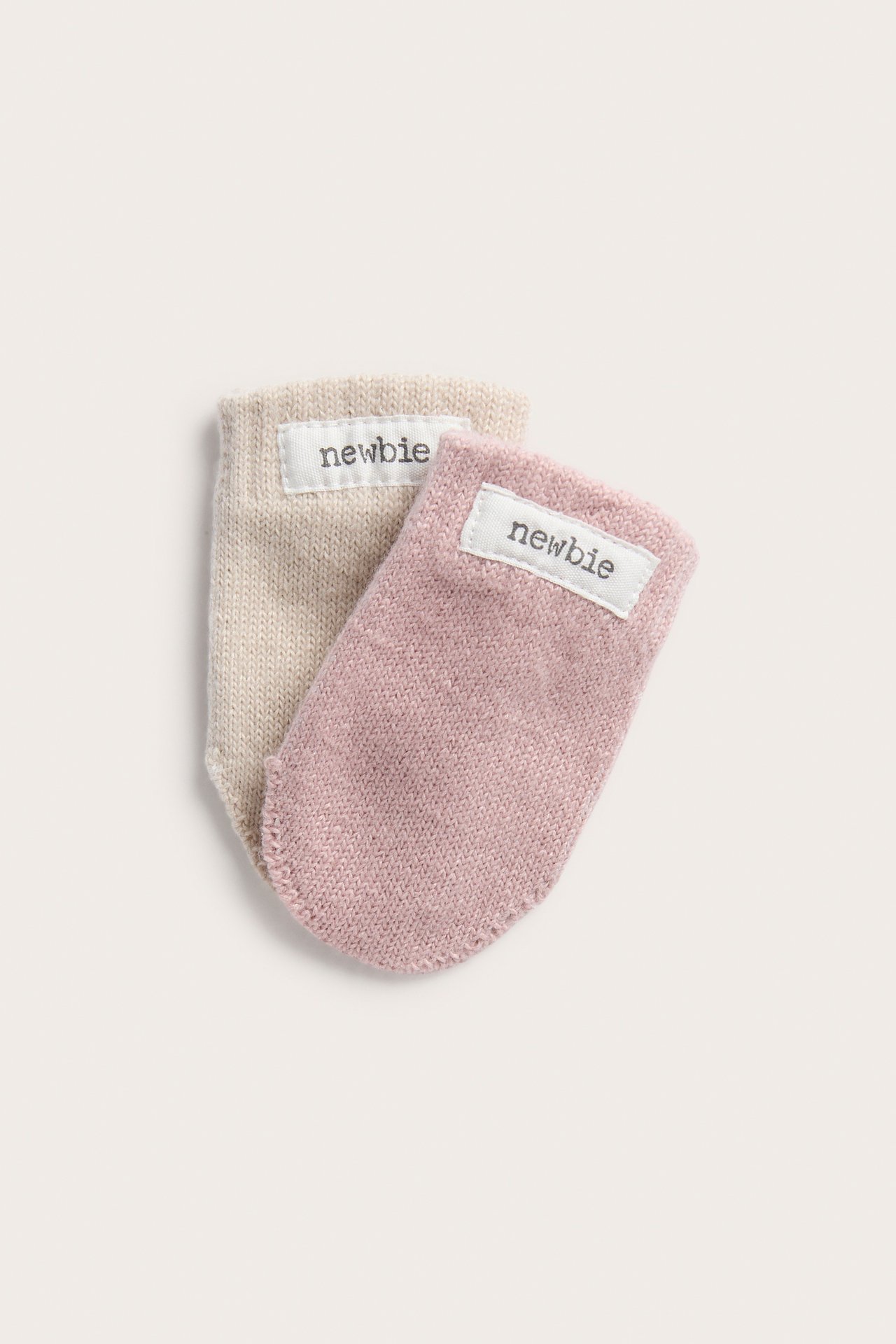 Nahaufnahme von zwei gestrickten Babyfäustlingen, einem rosa und einem beigen, mit 'newbie'-Etiketten.