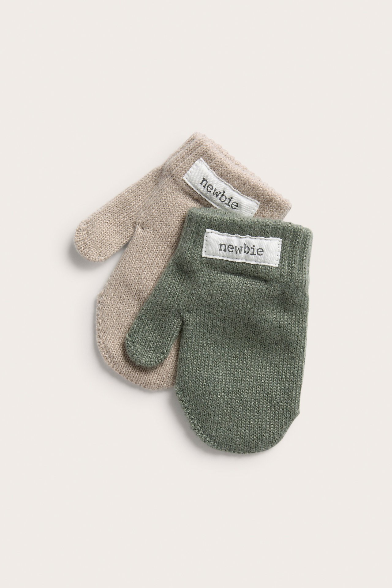 Nahaufnahme von zwei gestrickten Baby-Fäustlingen, einem beigen und einem grünen, mit 'newbie'-Etiketten.