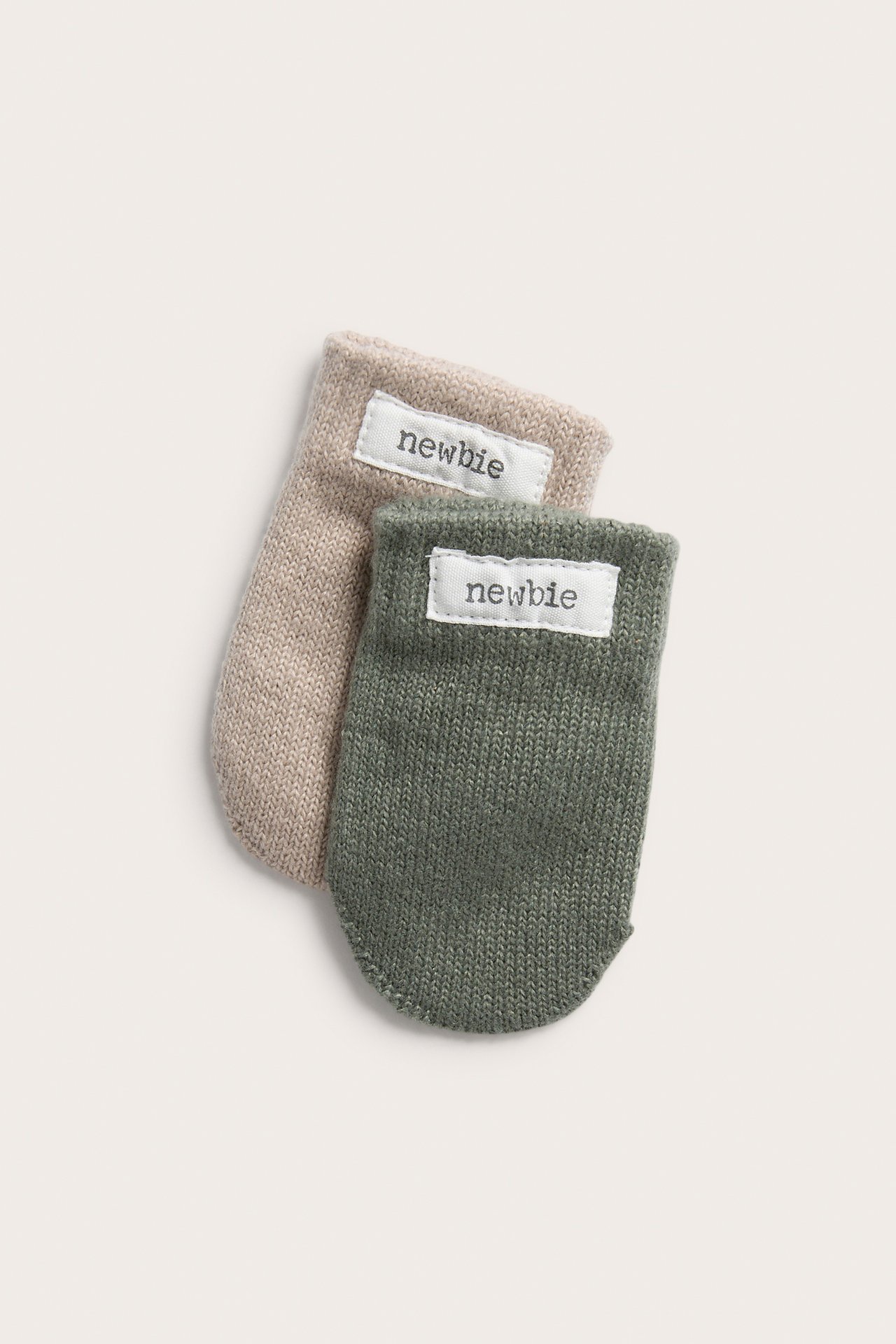 Nahaufnahme eines grünen gestrickten Babyhandschuhs mit Etikett, dahinter ein beiger gestrickter Babyhandschuh.