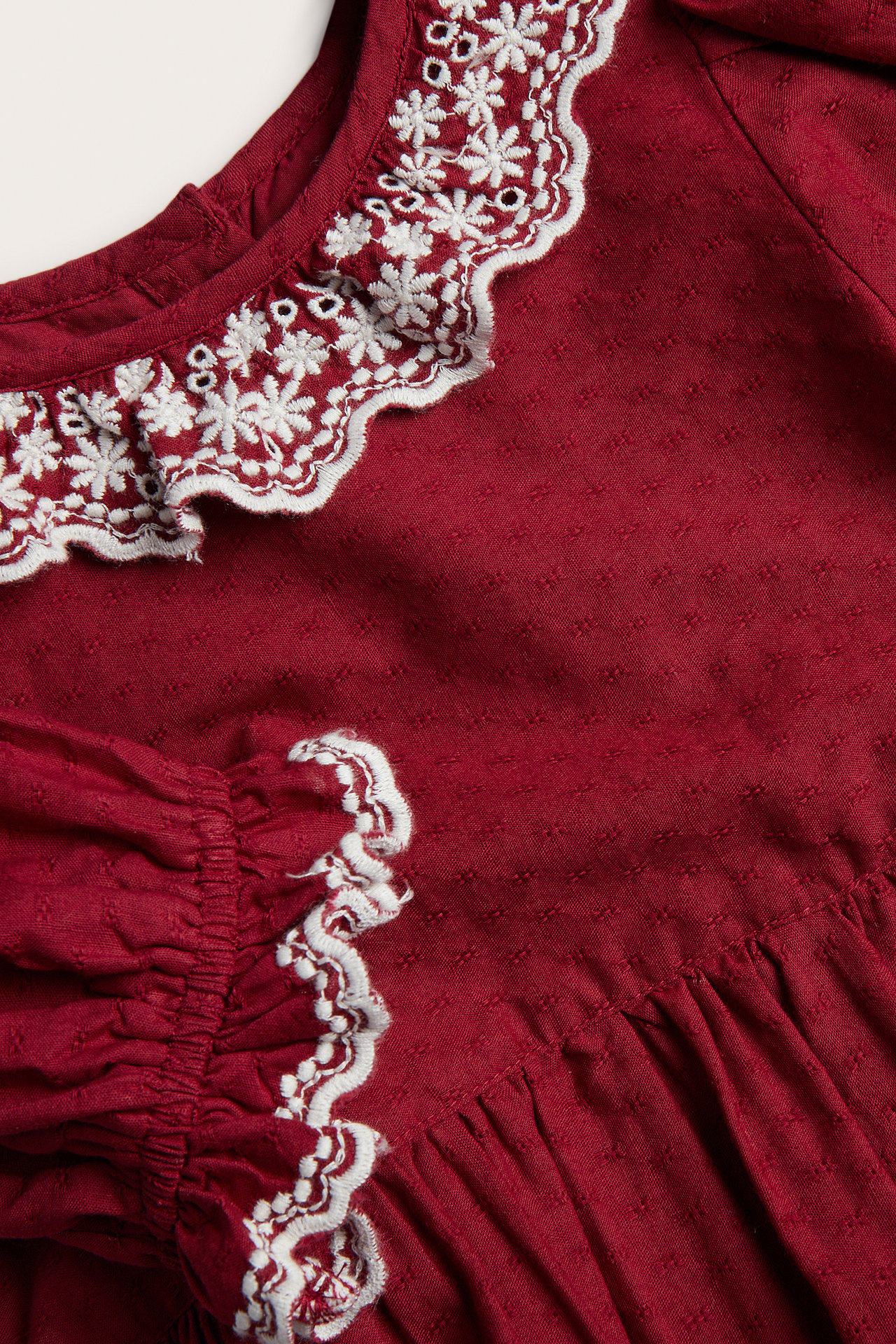 Close-up of a kid's burgundy dress with a white embroidered scalloped collar and sleeve trim.