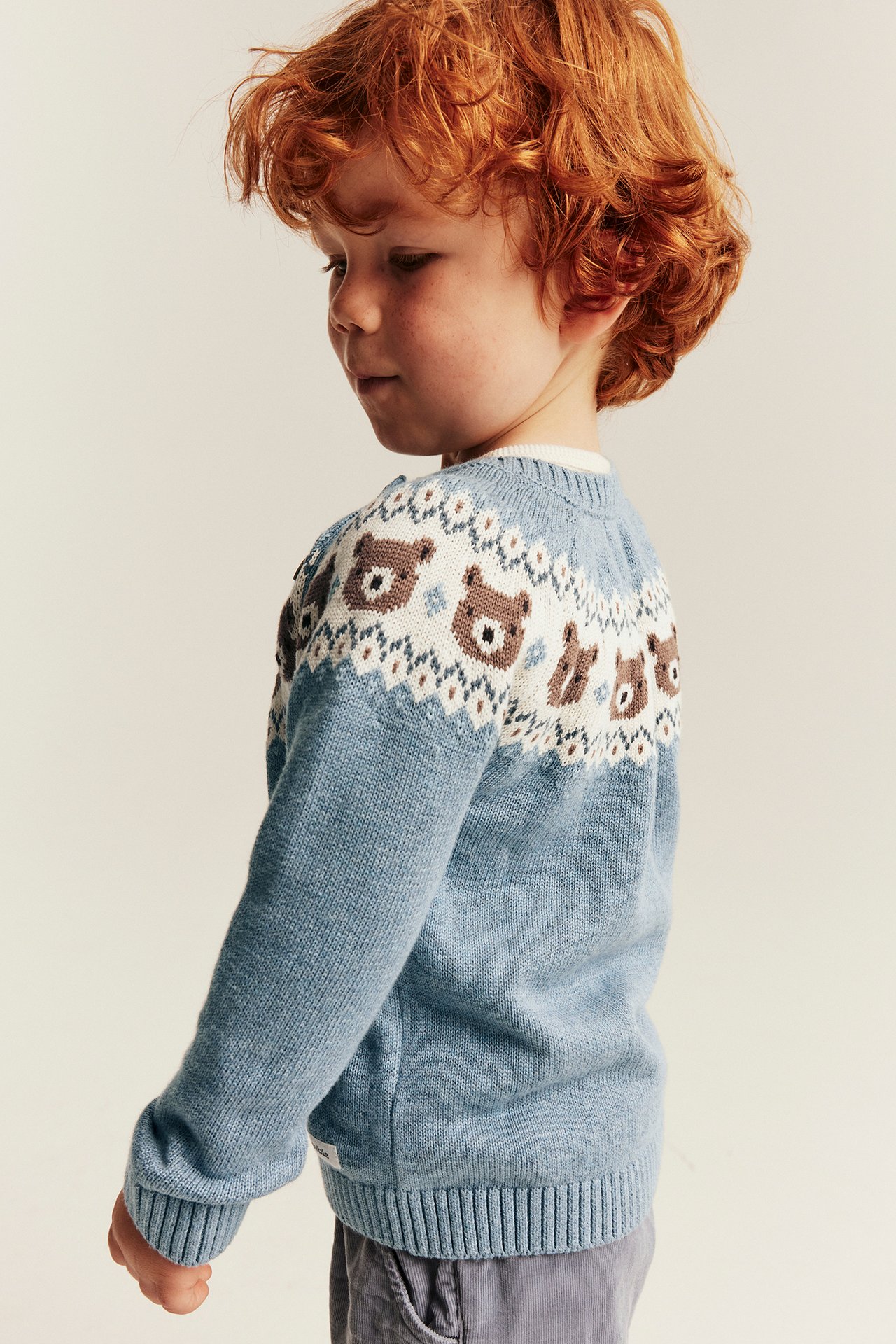 Seitenansicht eines Kindes in einem hellblauen Strickpullover mit Bärenmuster am Joch und einer grauen Cordhose.