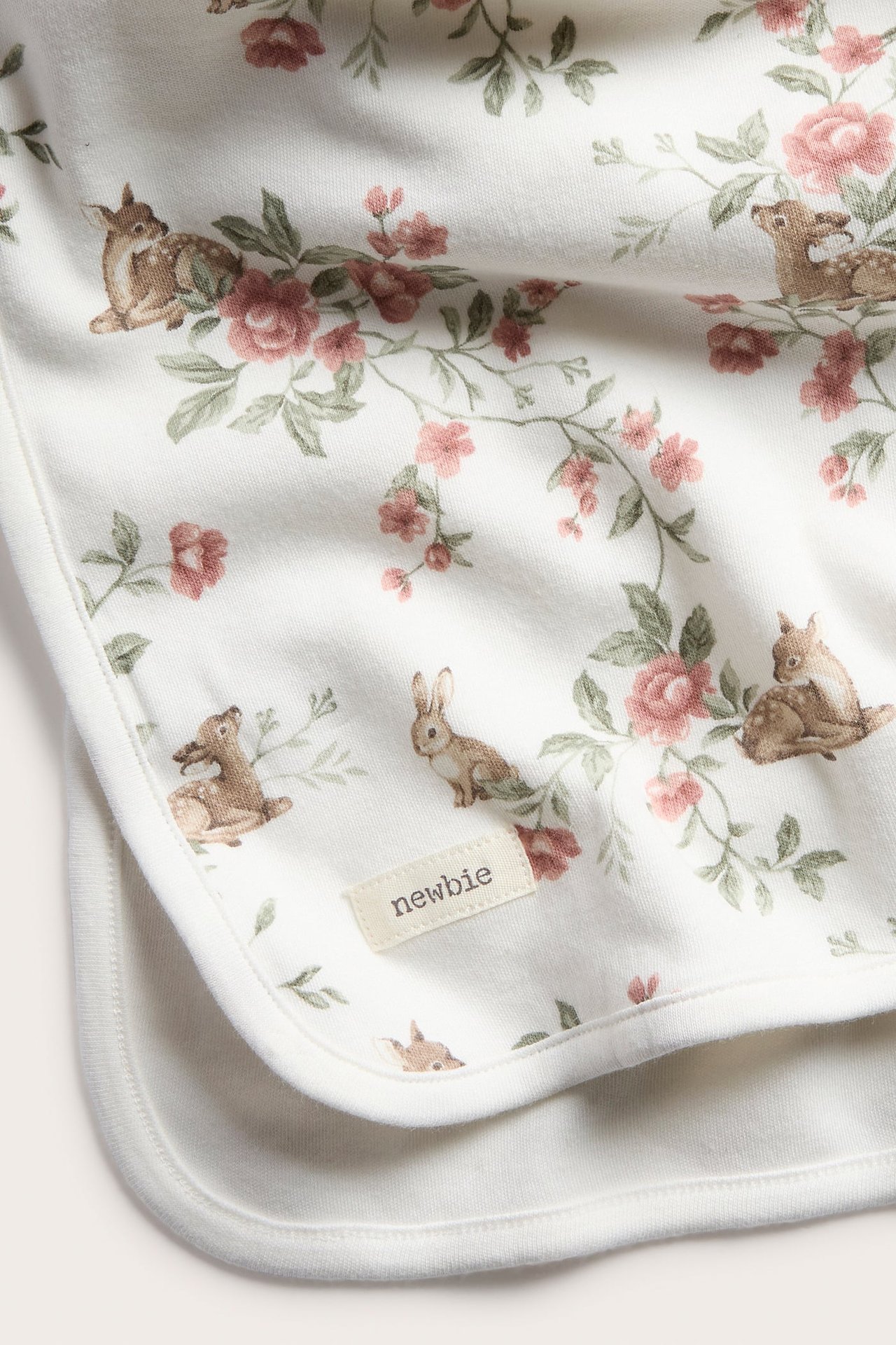 Close-up of an off-white cotton baby blanket with pink floral and fawn print, featuring a 'newbie' label.