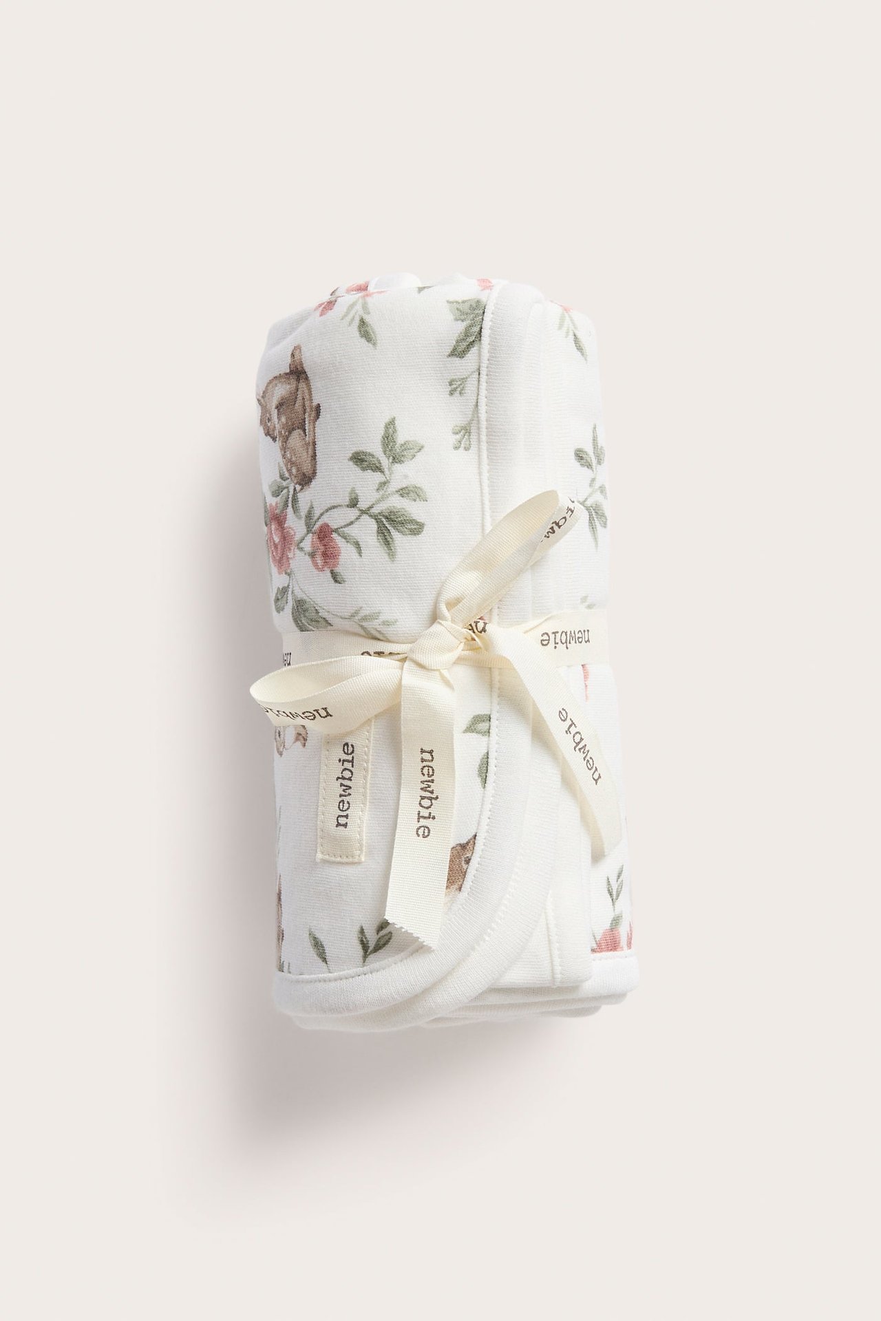 Front view of a rolled-up off-white cotton baby blanket with deer and floral print, tied with ribbon.